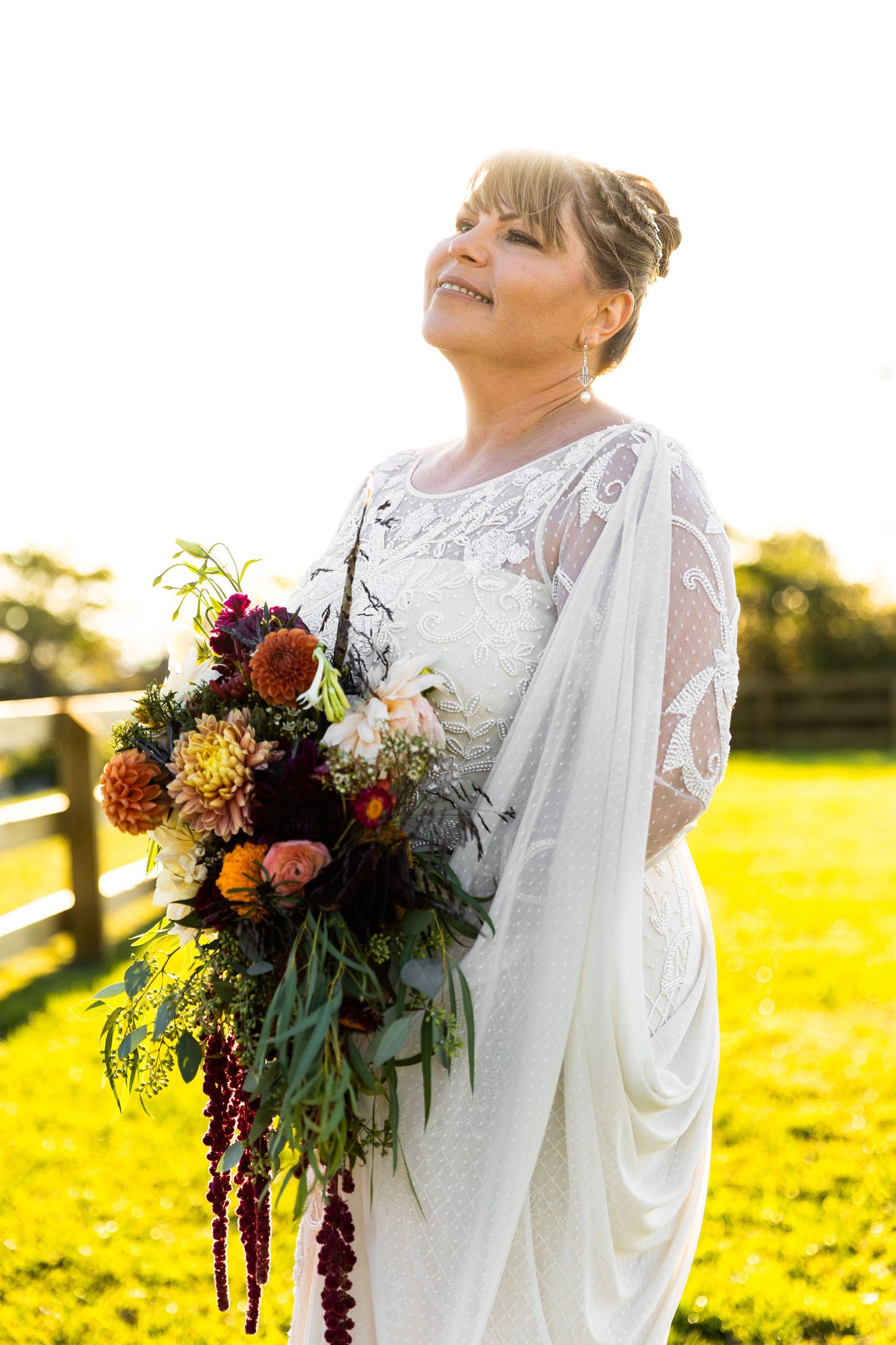Bride in beaded white gown holding lush colorful bouquet of dahlias smiling in golden sunlight.
