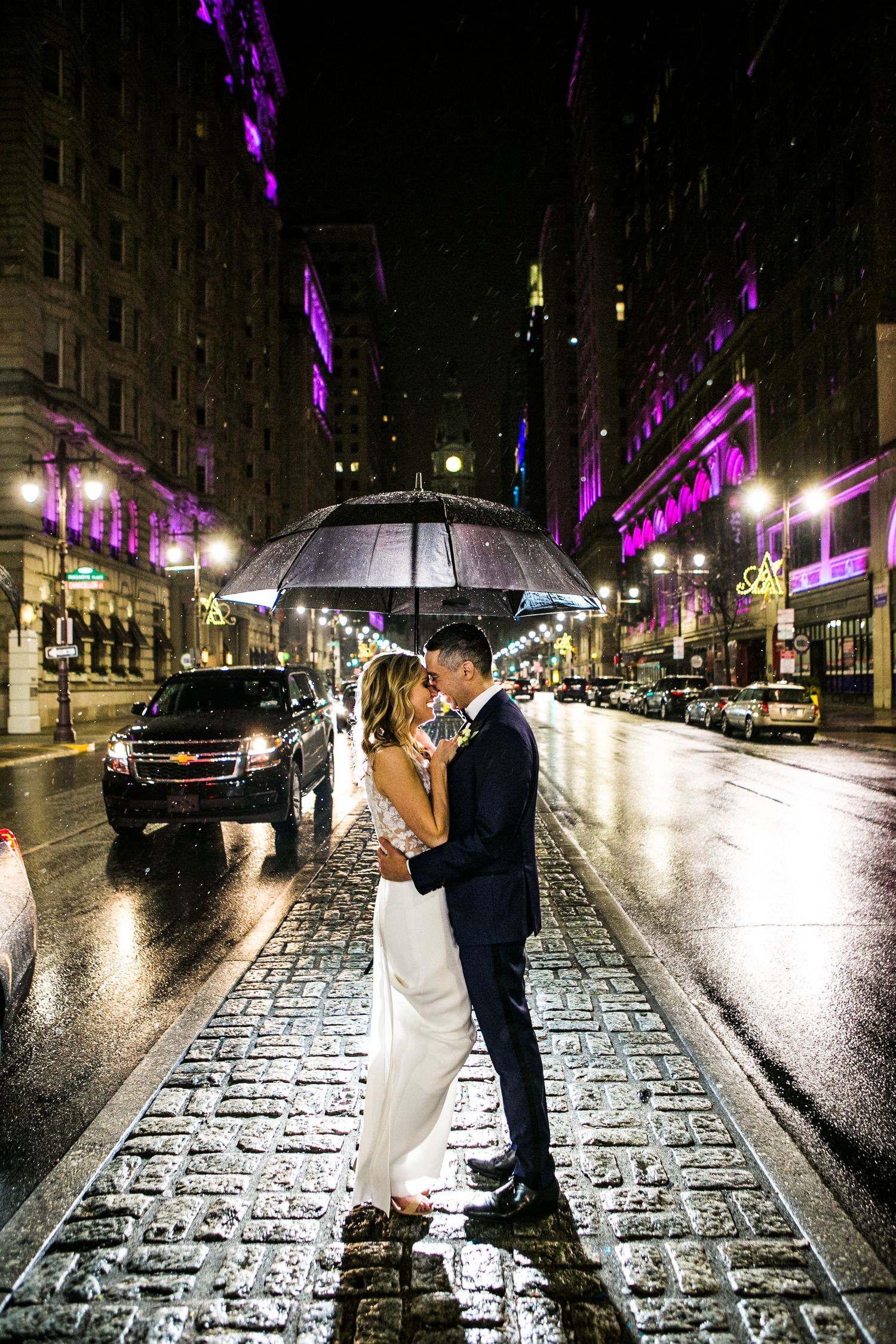 Bride and groom sharing a romantic kiss under a clear umbrella on a rainy city street at night.