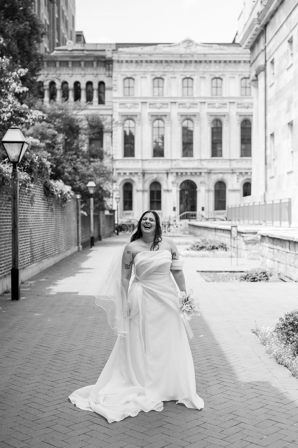 Black and white photo of laughing bride in flowing white gown with veil on brick path outdoors.