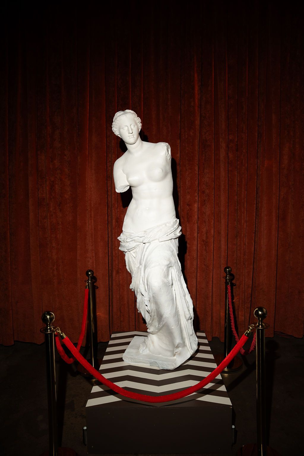 Classic Venus de Milo white marble replica statue displayed behind red velvet rope barriers on a checkered platform.
