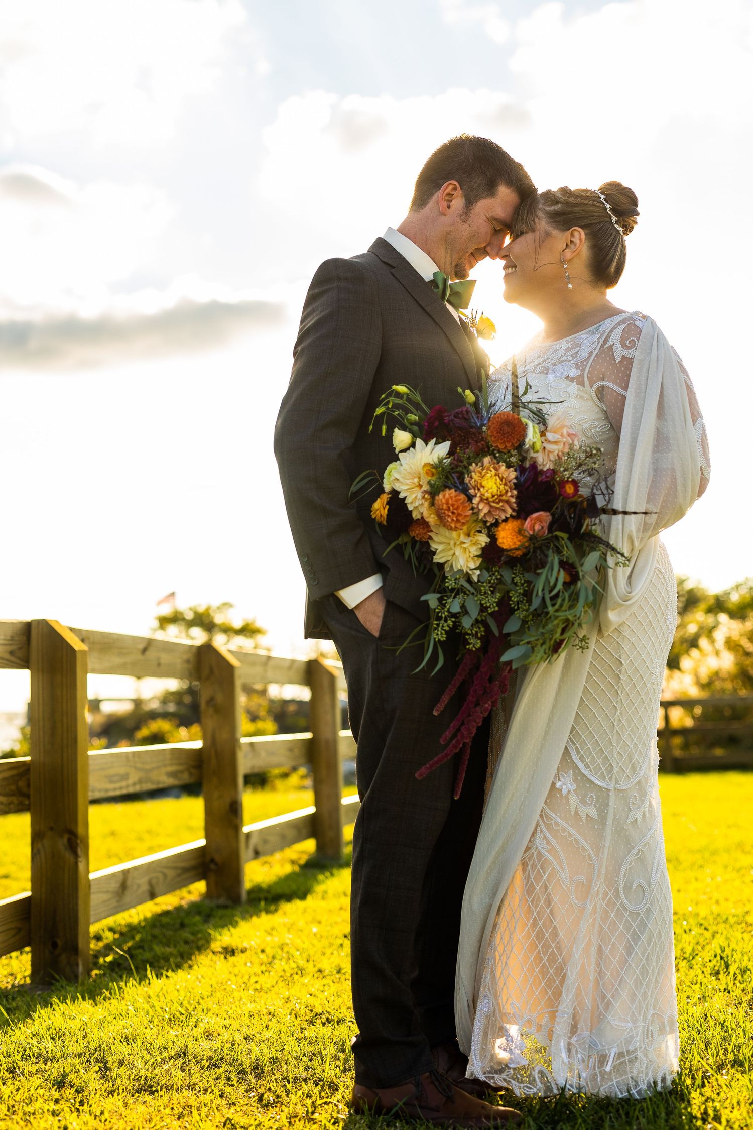 Bride and groom touch foreheads tenderly in golden sunset light beside rustic wooden farm fence.