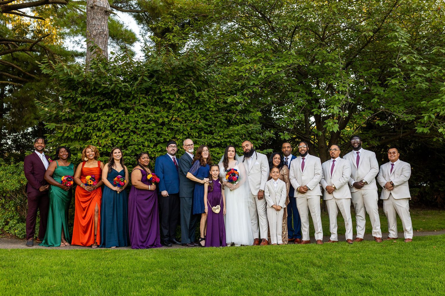 Large wedding party in colorful attire poses together on a lush green lawn surrounded by trees.