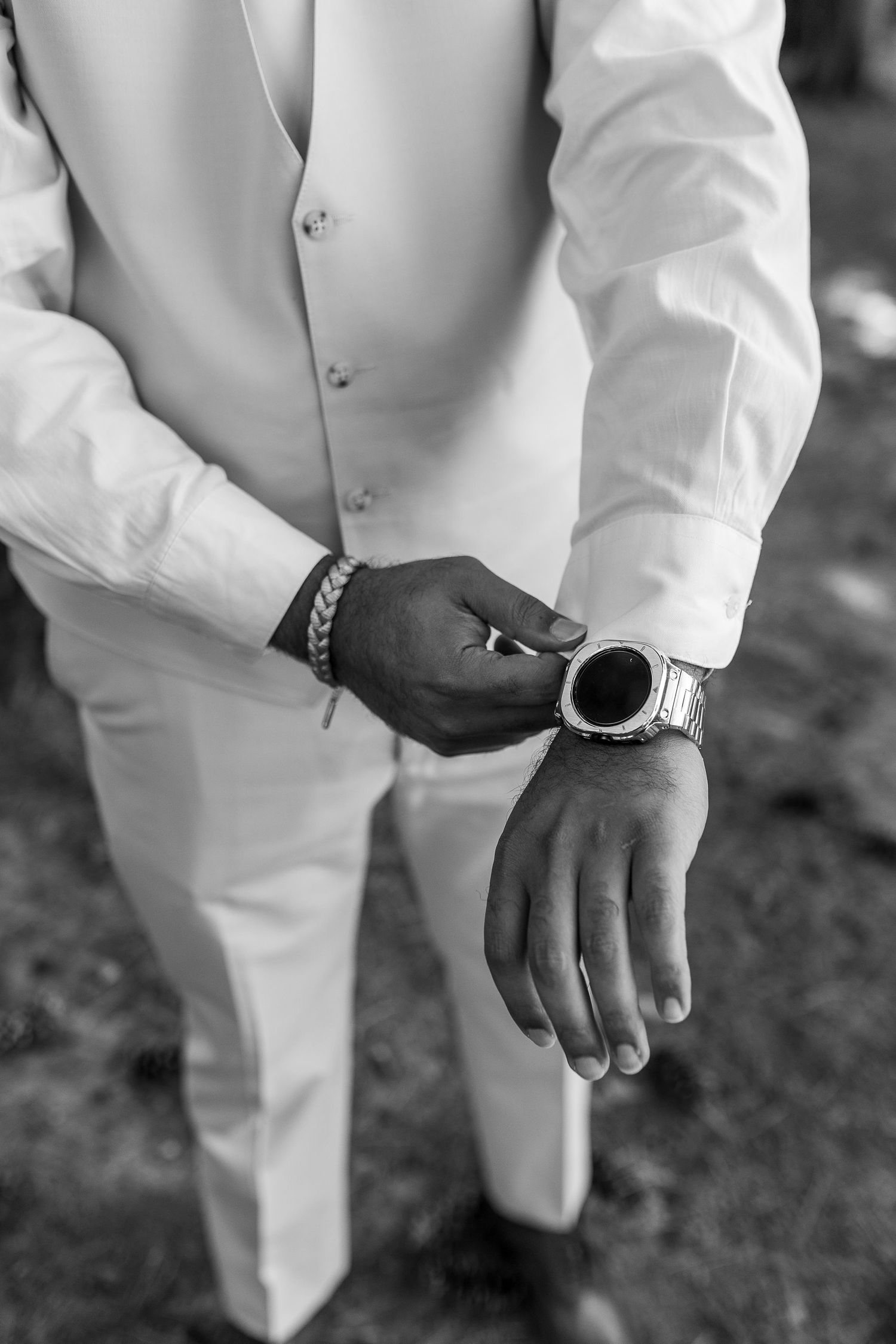 Black and white close-up of a groom in a white suit adjusting his watch with a bracelet on his wrist.