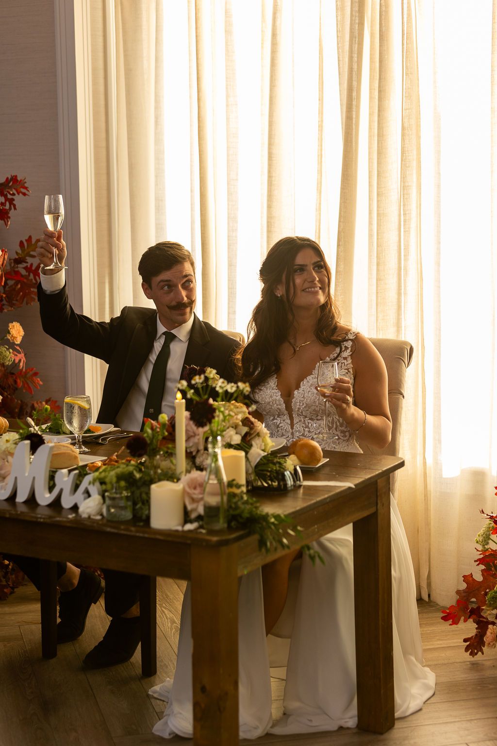 Bride and groom raise champagne glasses for toast at floral decorated sweetheart table at wedding.