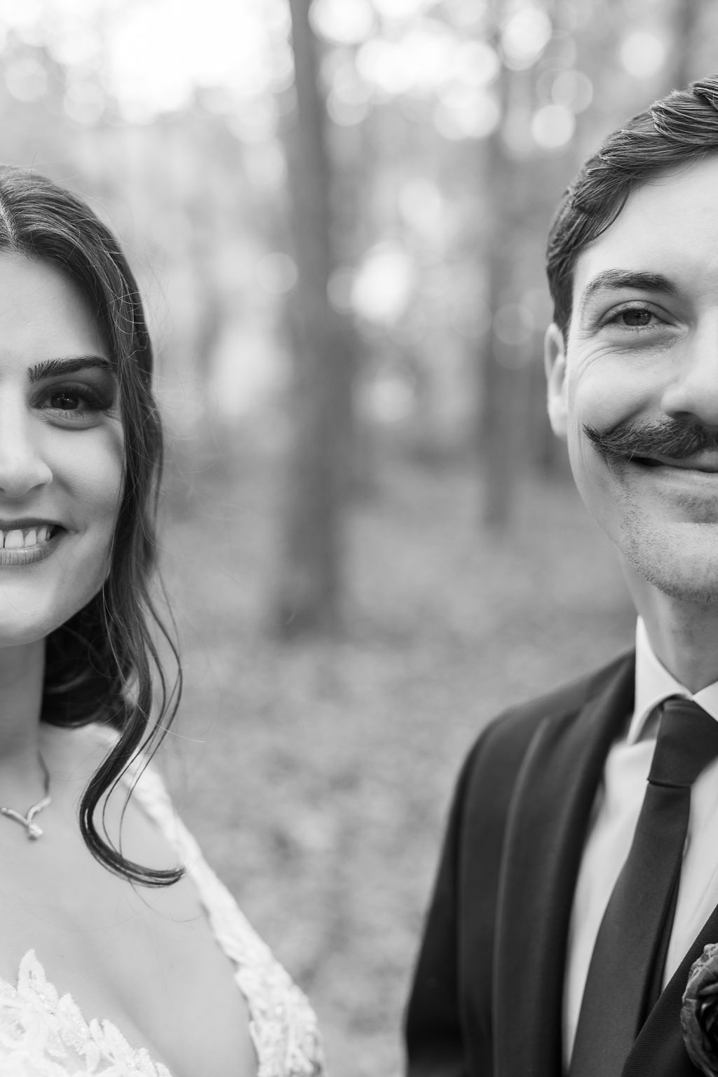 Black and white close-up portrait of smiling bride and mustachioed groom in wedding attire outdoors.