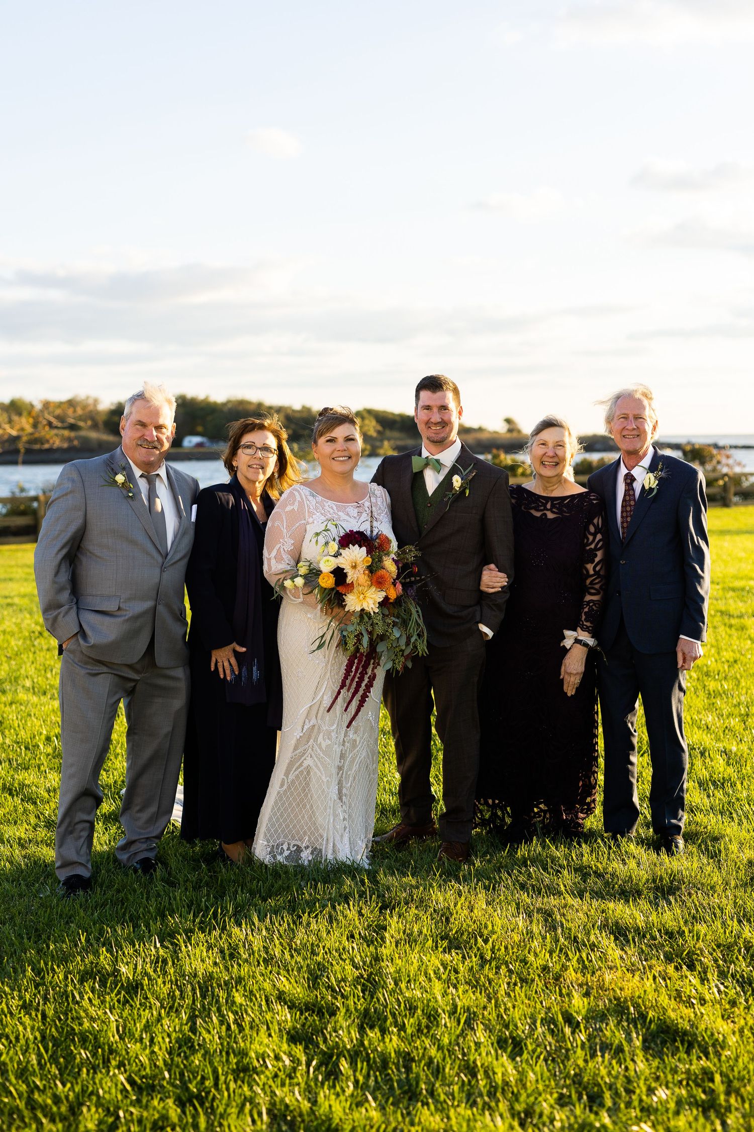 Wedding party group portrait of bride, groom, and four parents on a scenic waterfront lawn.