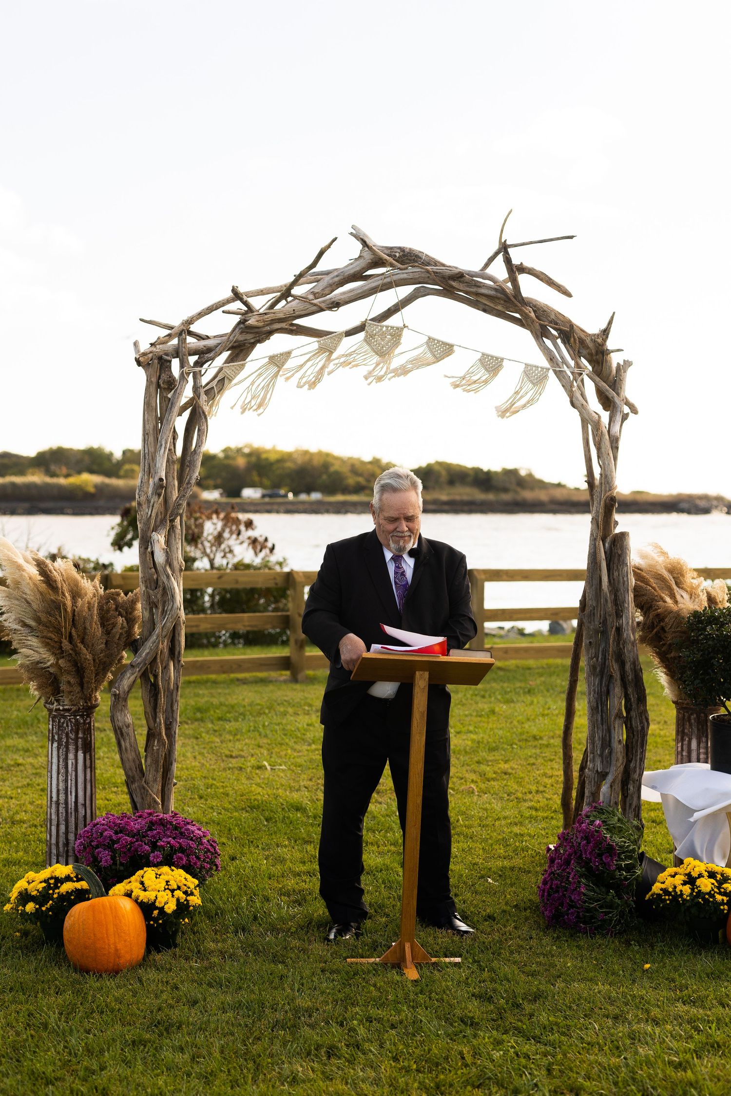 Officiant reads ceremony script at rustic driftwood arch decorated with pumpkins by waterfront.