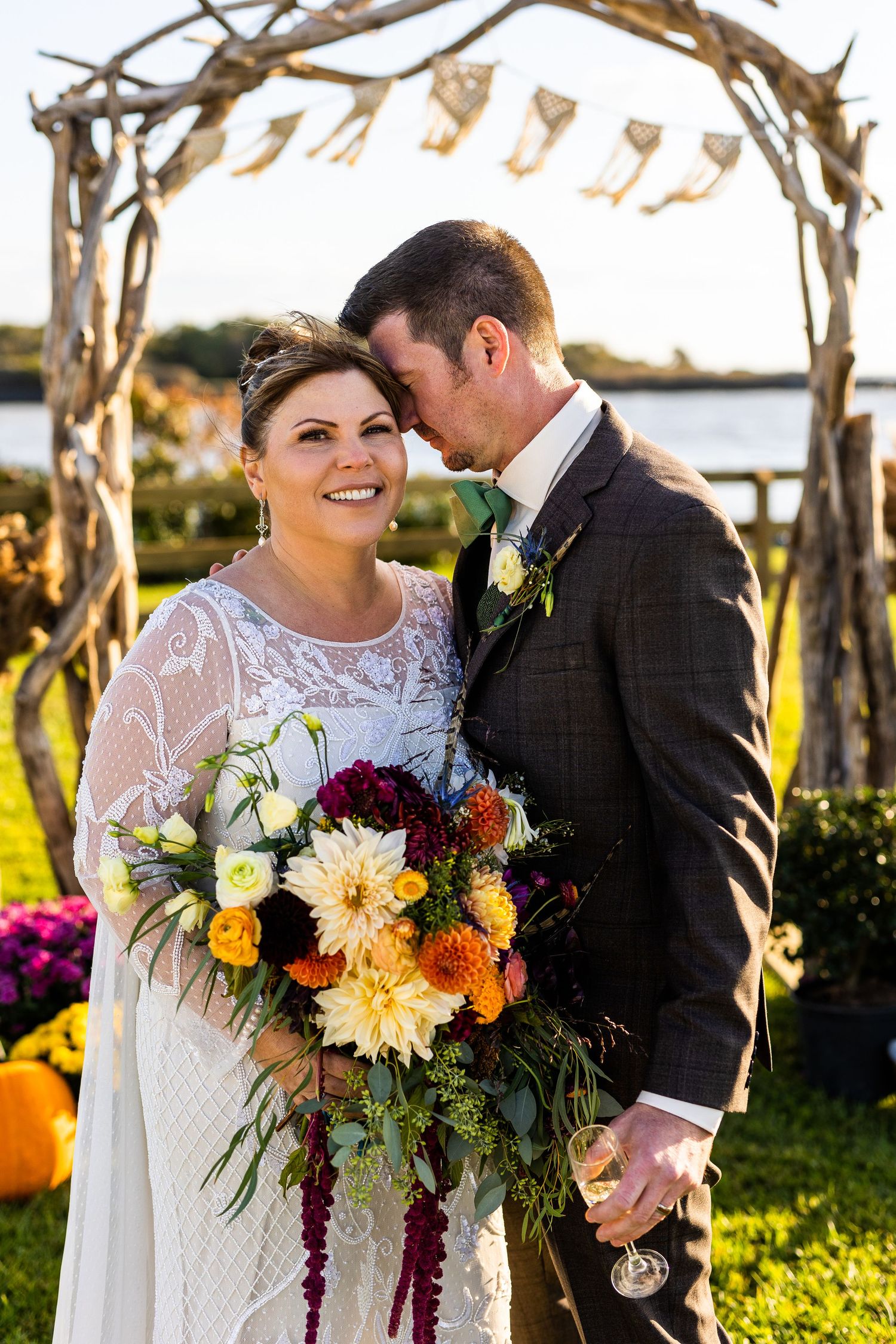 Bride smiles holding vibrant autumn bouquet as groom nuzzles her cheek under driftwood arch at fall wedding.