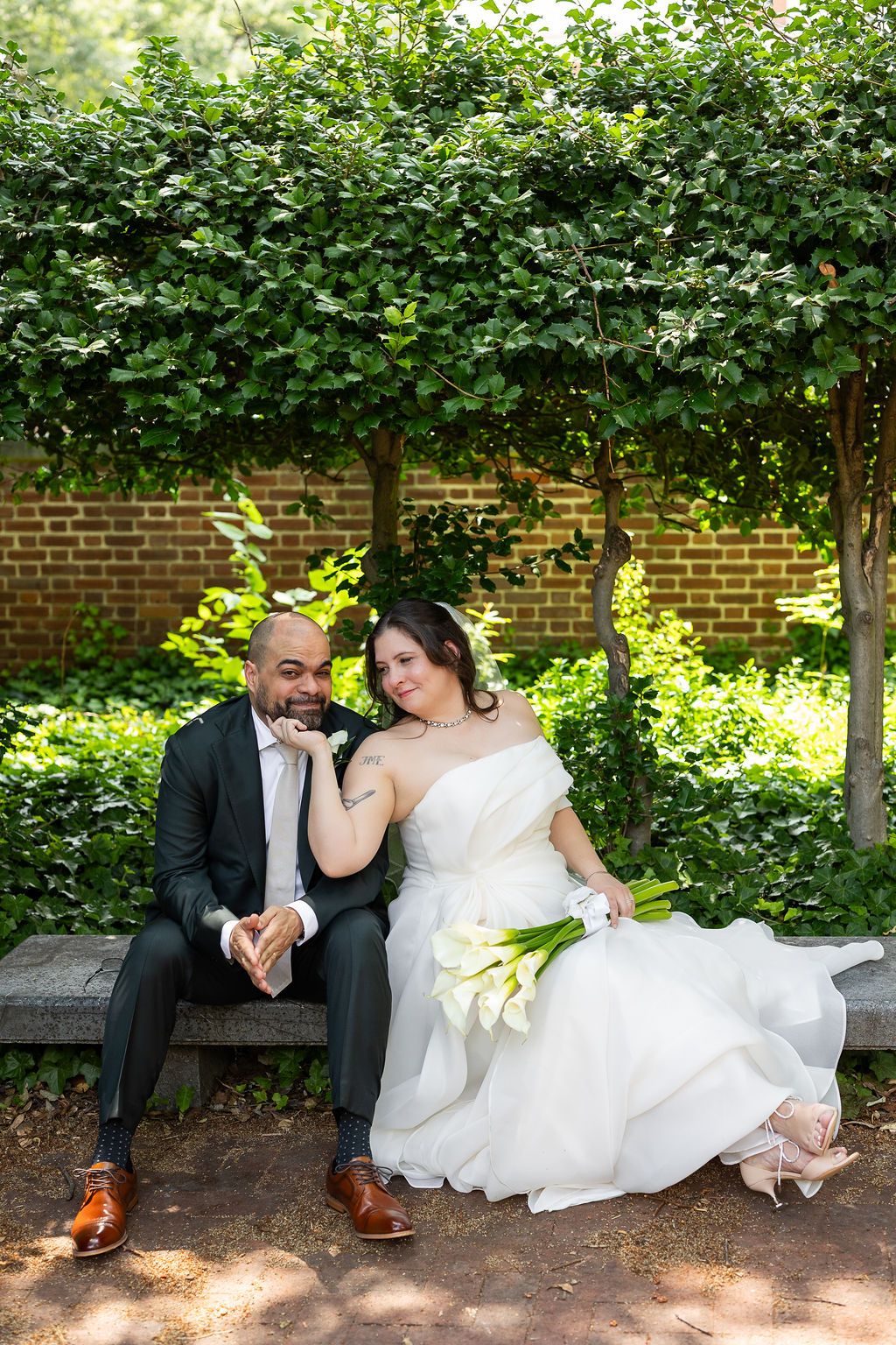 Bride resting head on groom's shoulder while seated on garden bench, holding calla lilies in front of brick wall.