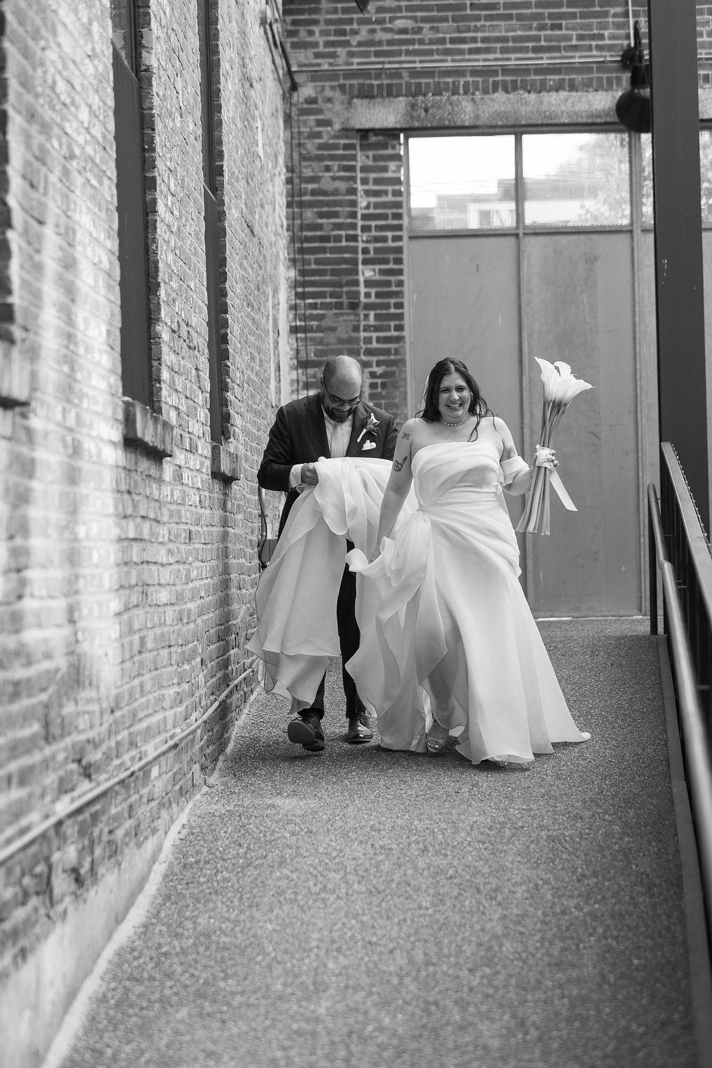 Black and white photo of groom helping bride with her dress in a narrow walkway.