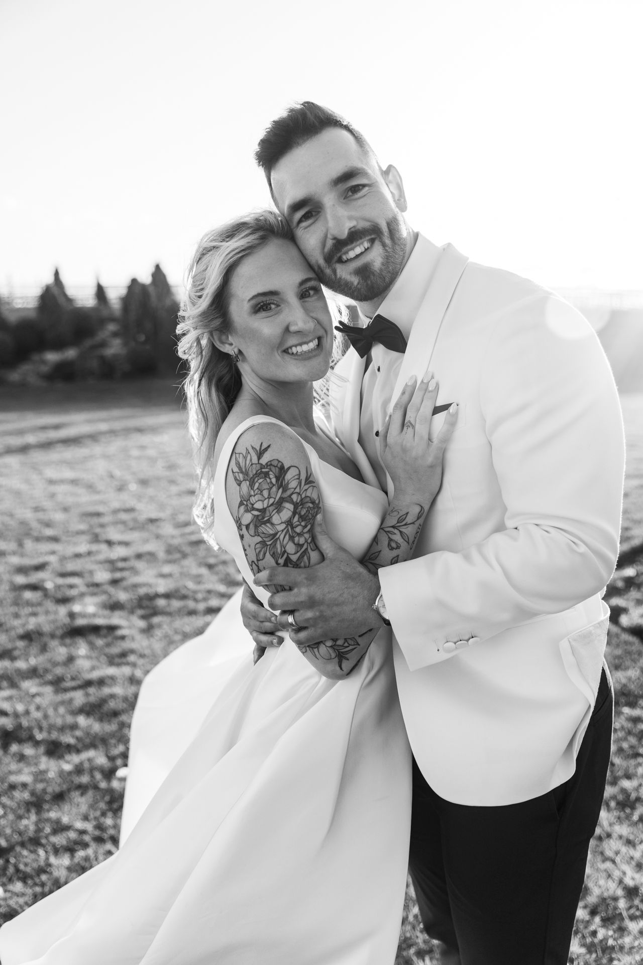 Black and white portrait of tattooed bride and groom embracing and smiling at outdoor wedding.