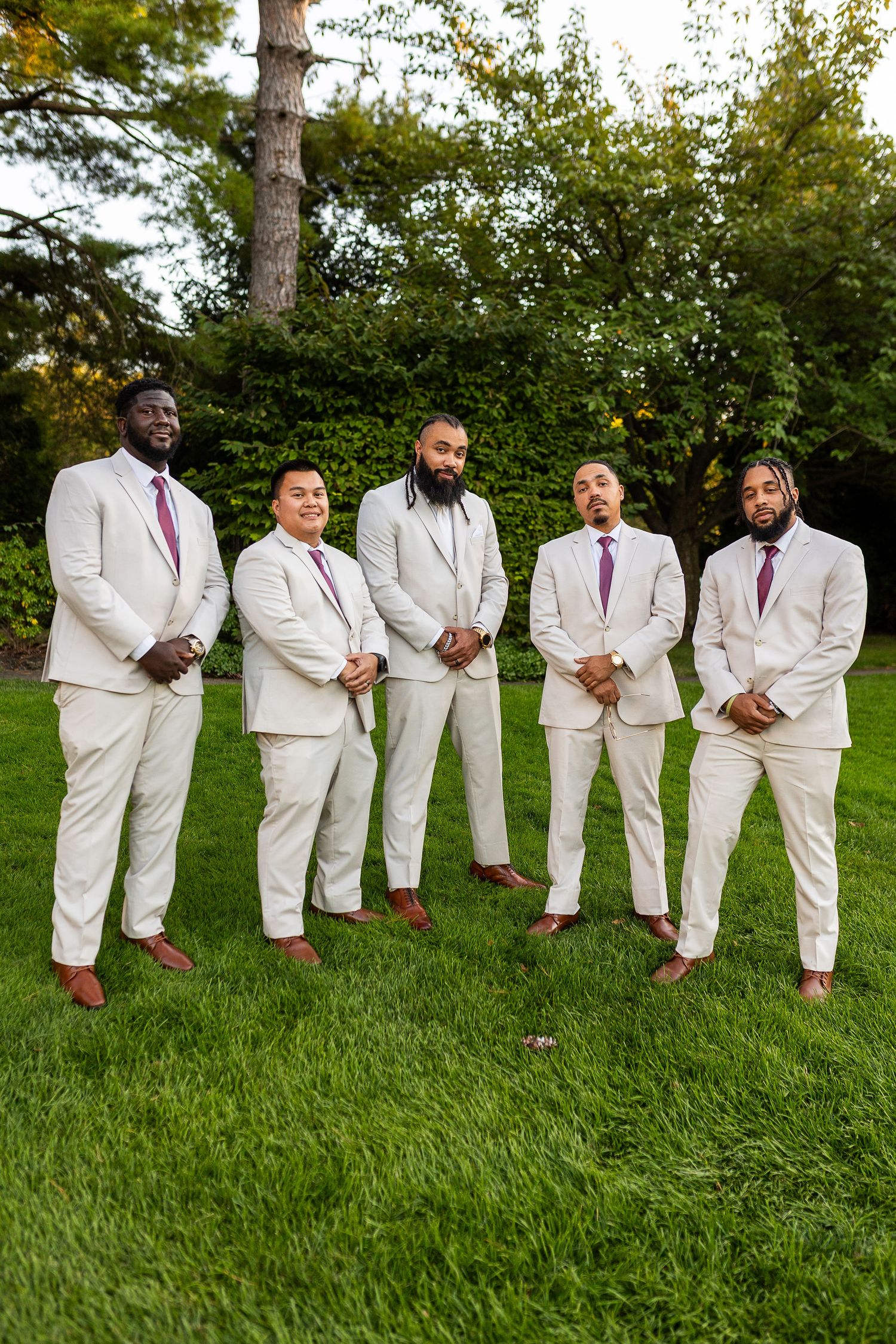 Five groomsmen in matching cream suits with purple ties pose confidently on a green lawn.