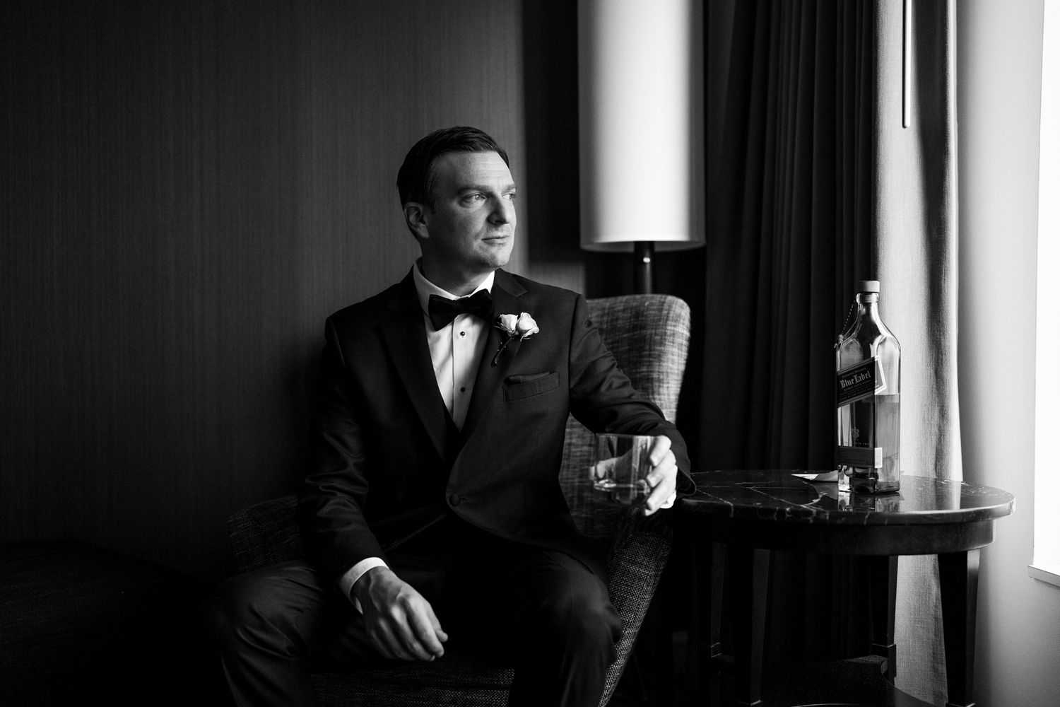Groom in black tuxedo sitting in a hotel chair holding a whiskey glass in a black and white photo.