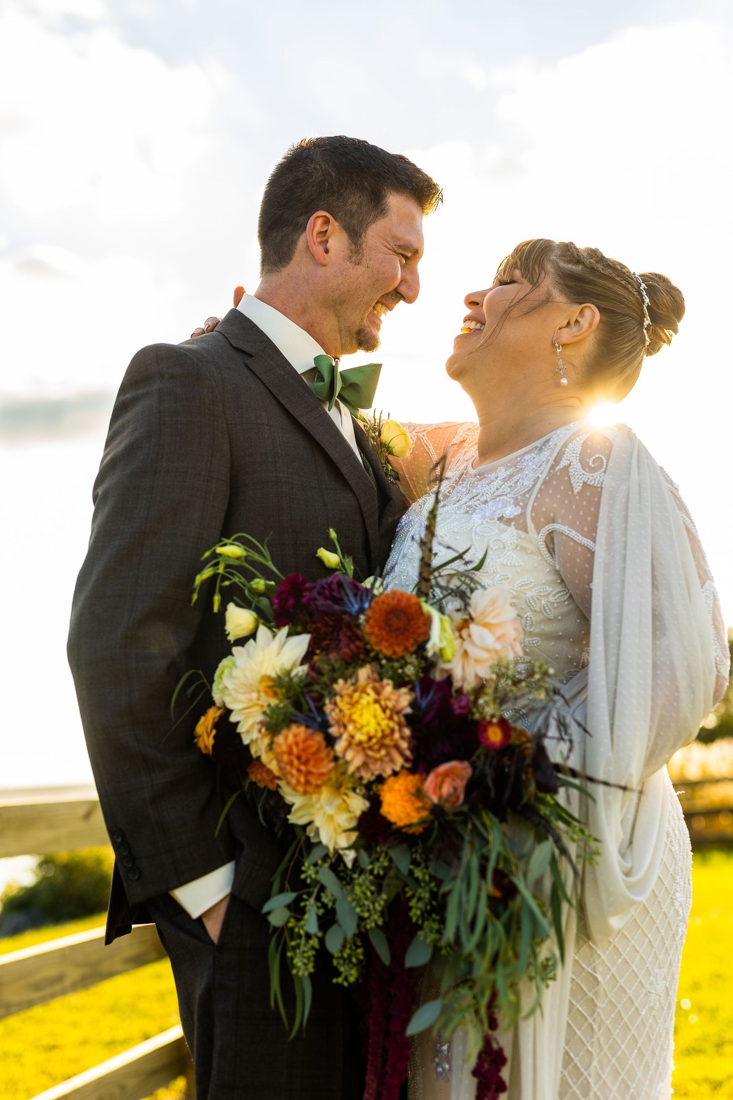Laughing bride and groom holding colorful fall bouquet with golden sunset backlight in outdoor setting.