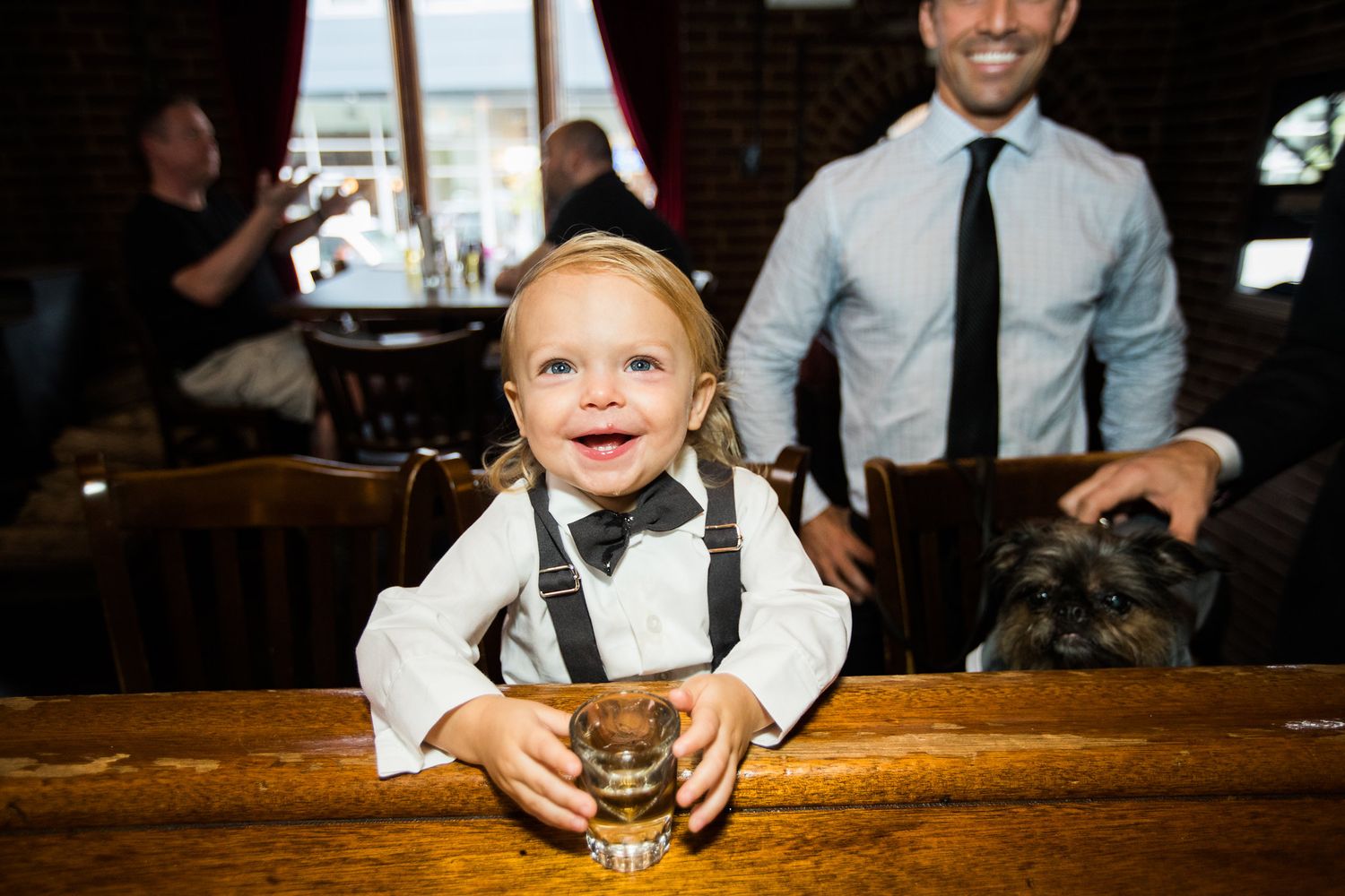 Adorable toddler ring bearer in suspenders and bow tie smiling at bar during wedding reception.