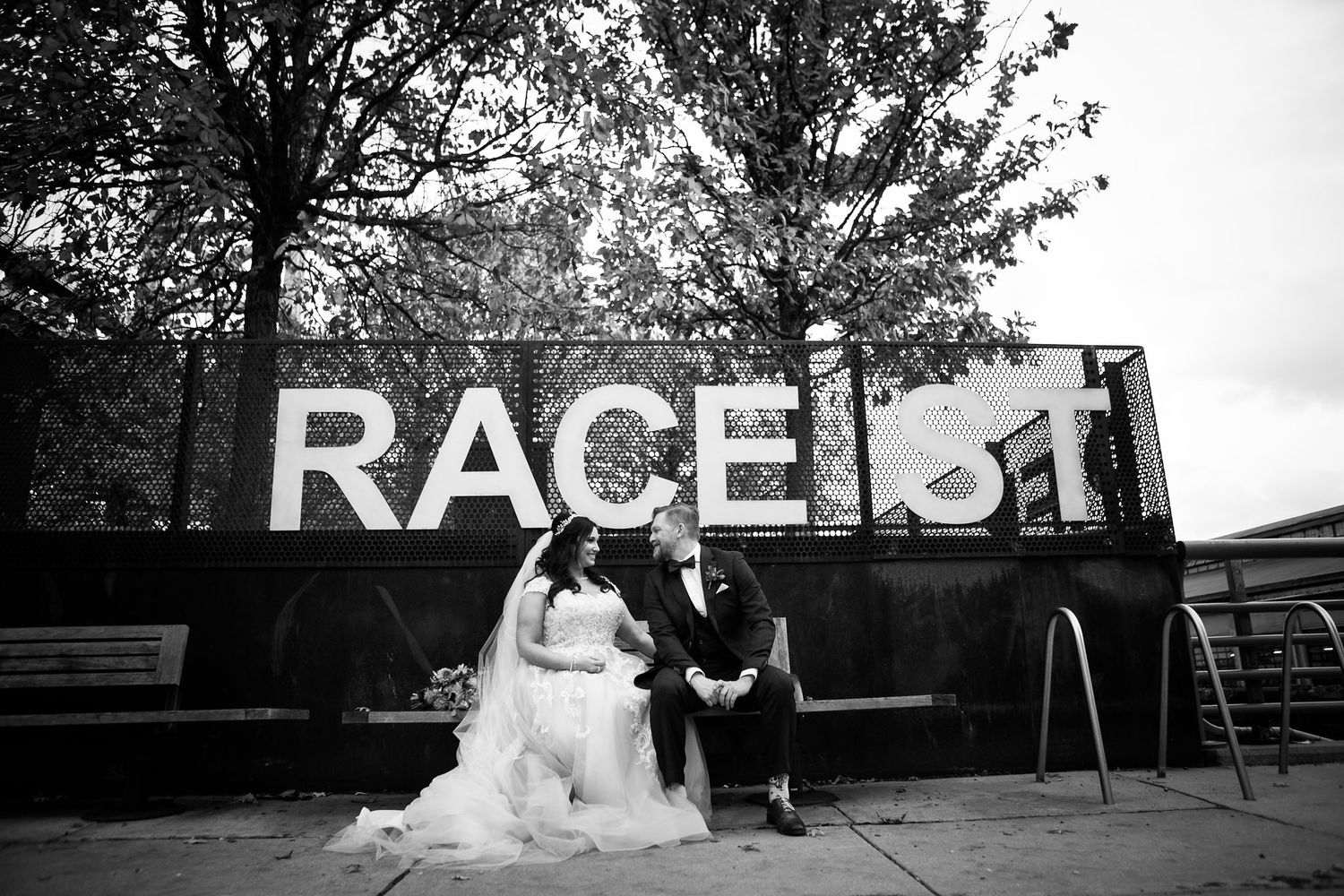 Black and white wide shot of bride and groom seated near Race Street sign surrounded by autumn trees.