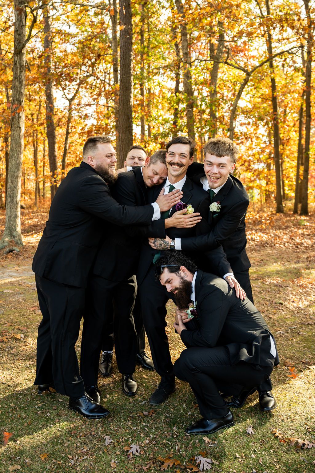 Groom in black suit laughing as groomsmen playfully embrace him among colorful fall trees.