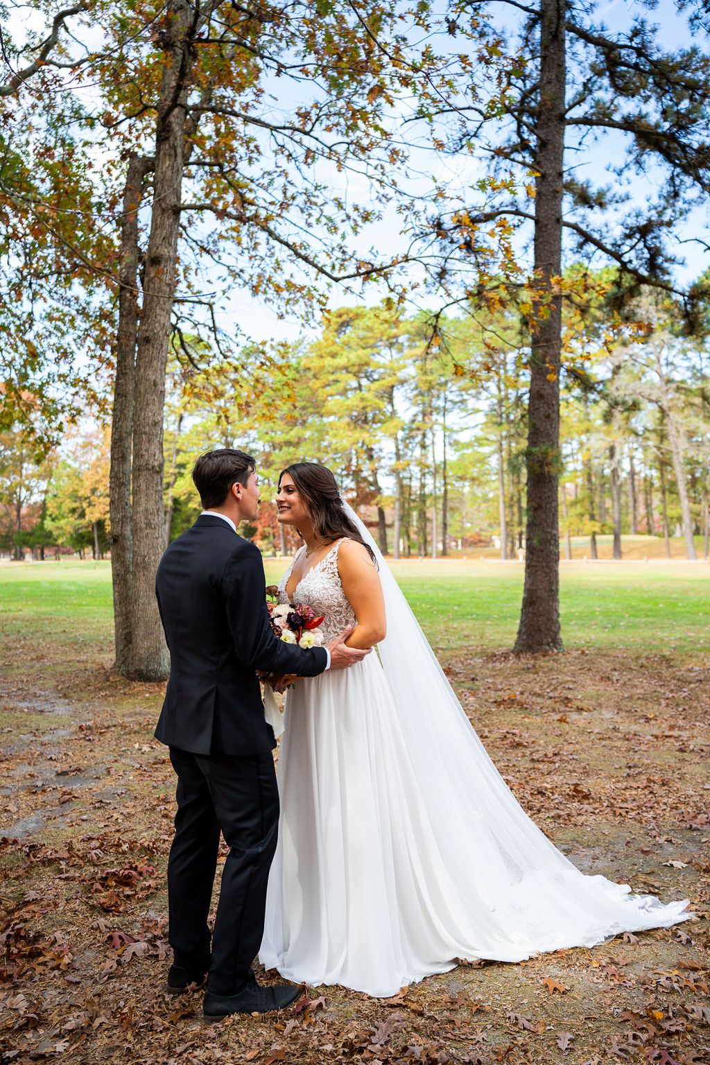 Bride and groom hold hands and smile at each other among tall pine trees in fall foliage.