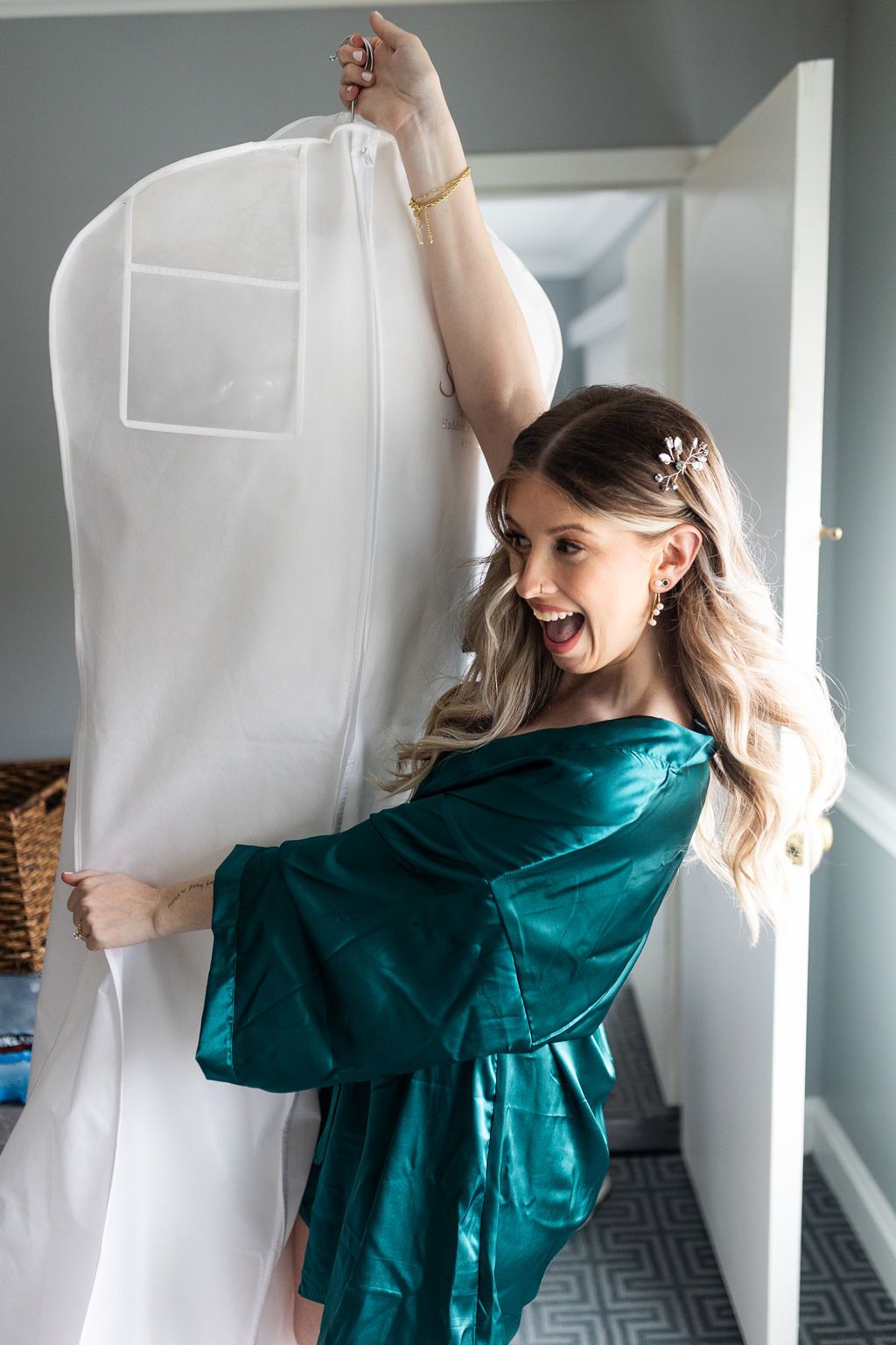 Excited bridesmaid in teal satin robe holding up the bride's wedding dress in a garment bag.
