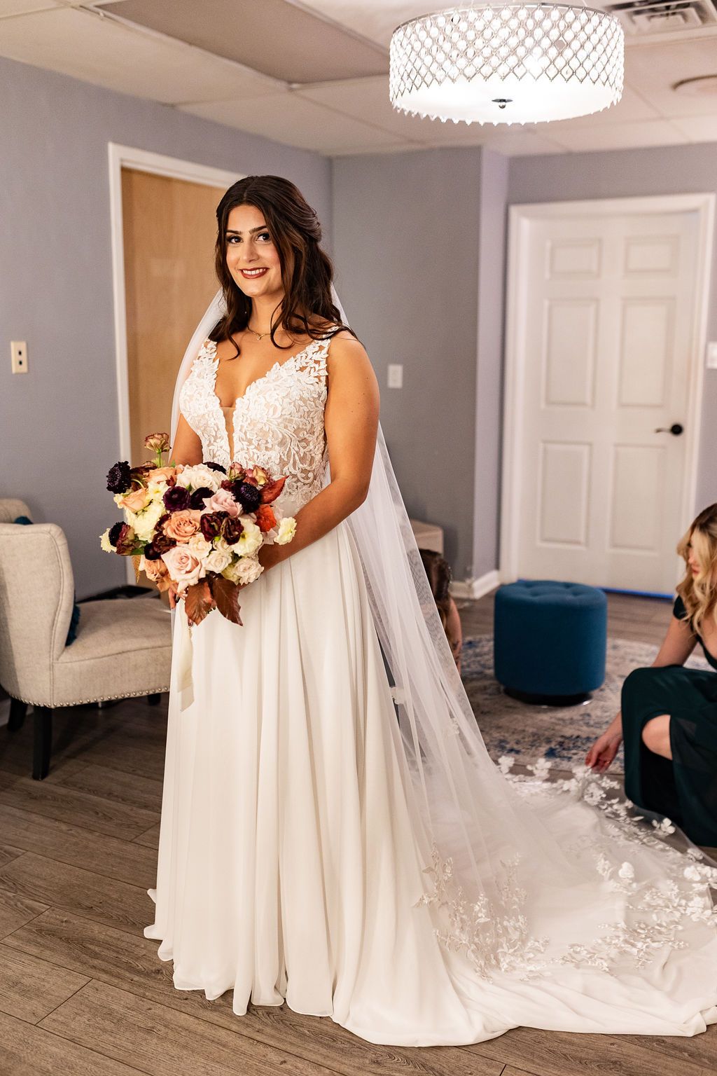 Bride in a lace bodice wedding gown with veil holding an autumn bouquet of burgundy and blush roses in a bridal suite.