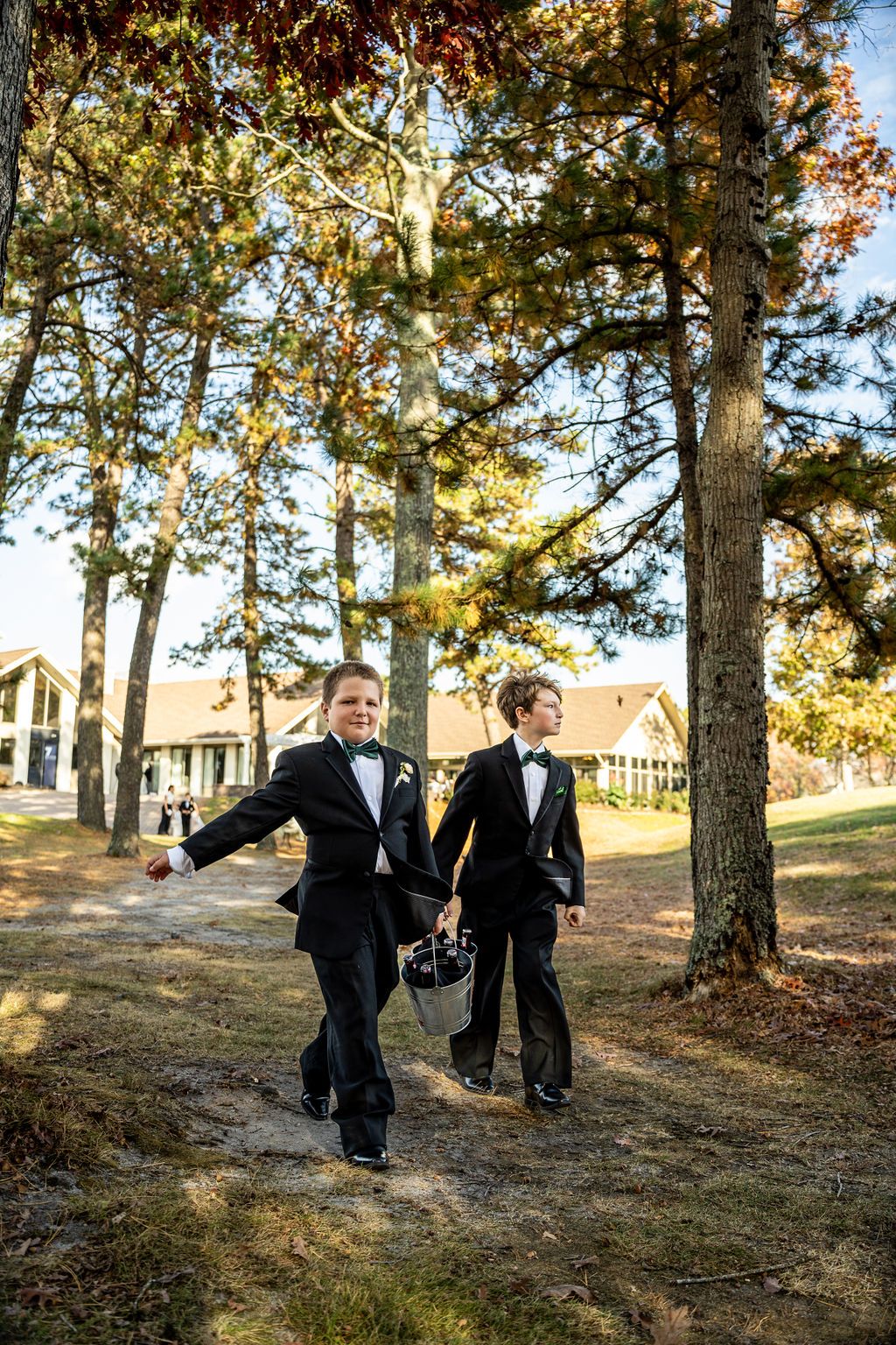 Two young ring bearers in black tuxedos walk down outdoor wedding path among tall pine trees on sunny day.