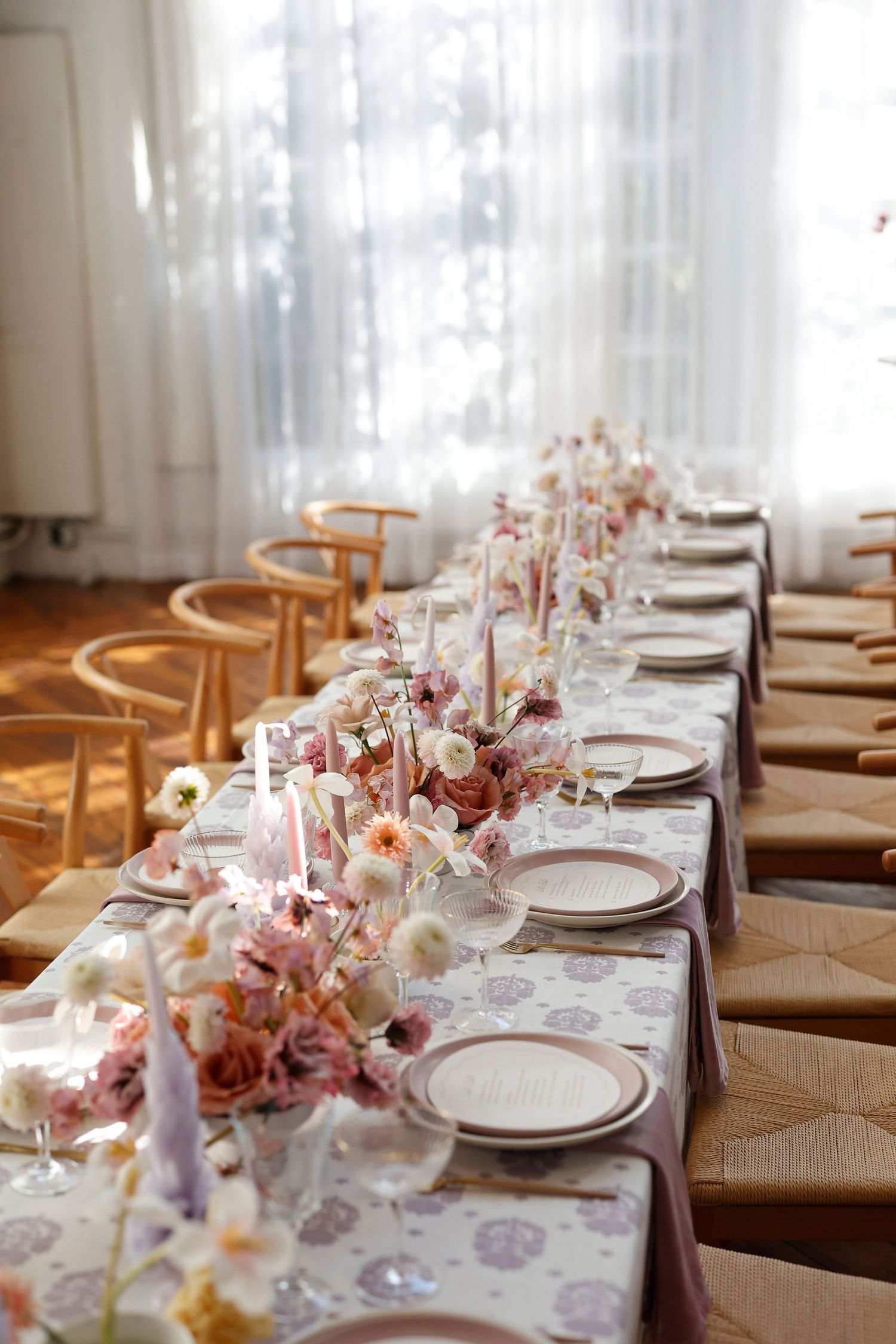 Sunlit wedding reception long table adorned with pink floral centerpieces, taper candles, and patterned linens.