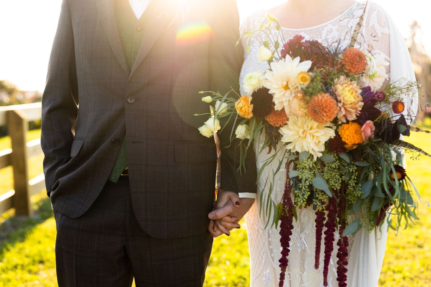 Close-up of bride and groom holding hands with vibrant floral bouquet at sunset.