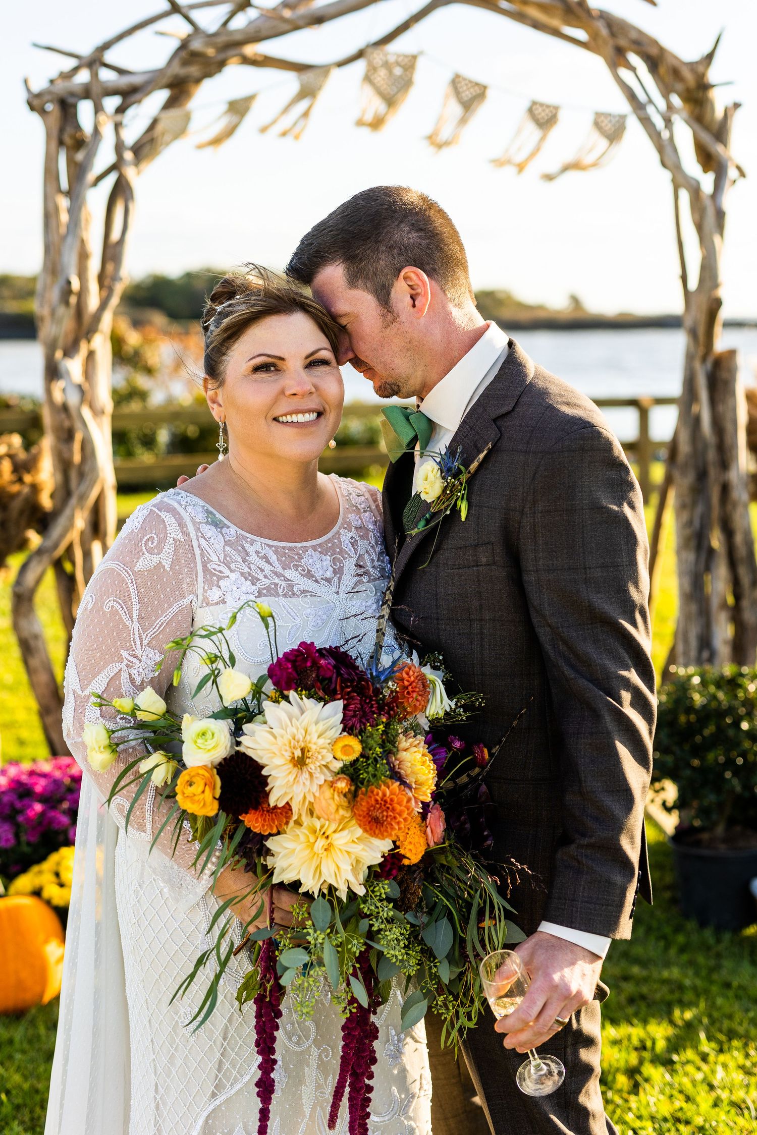 Bride and groom embracing under rustic wooden arch with colorful fall bouquet and water view behind them.