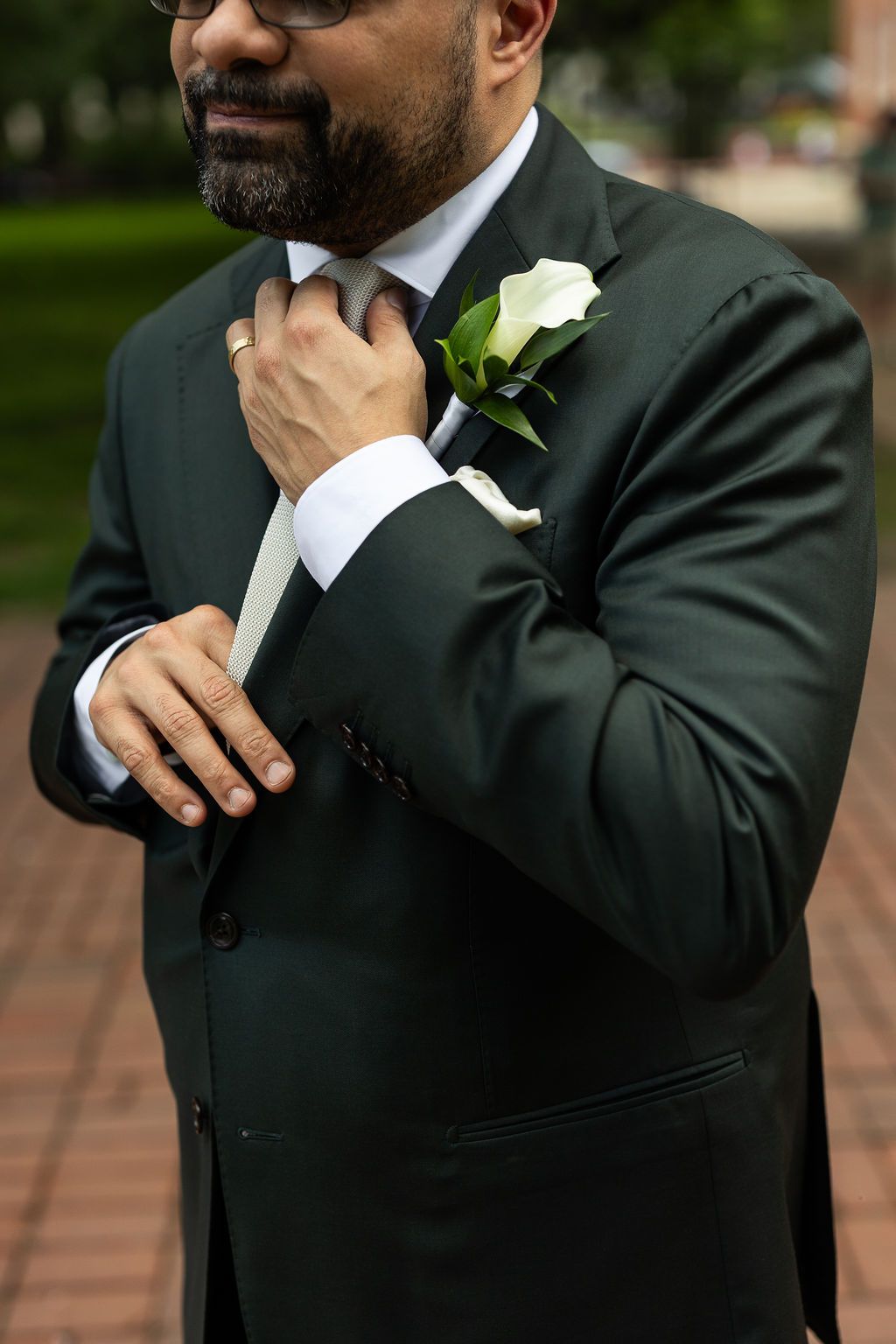 Close-up of groom in dark suit adjusting his tie with a white calla lily boutonniere on lapel.