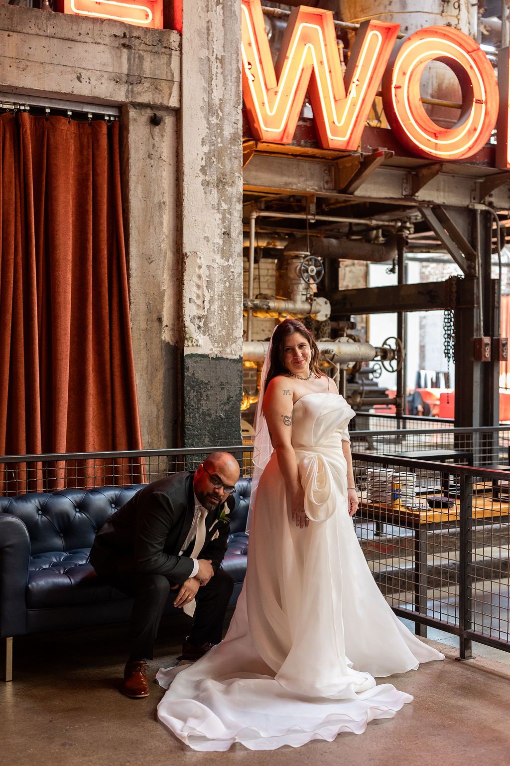 Groom adjusting brides white wedding gown train while posing under glowing neon signs in industrial venue.