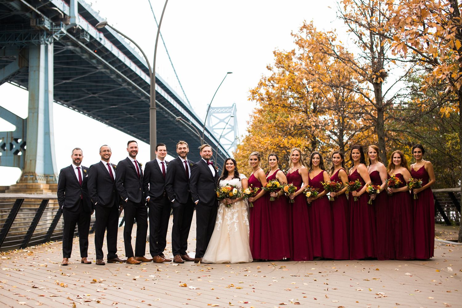 Wedding party in burgundy and black attire poses under autumn trees near suspension bridge.
