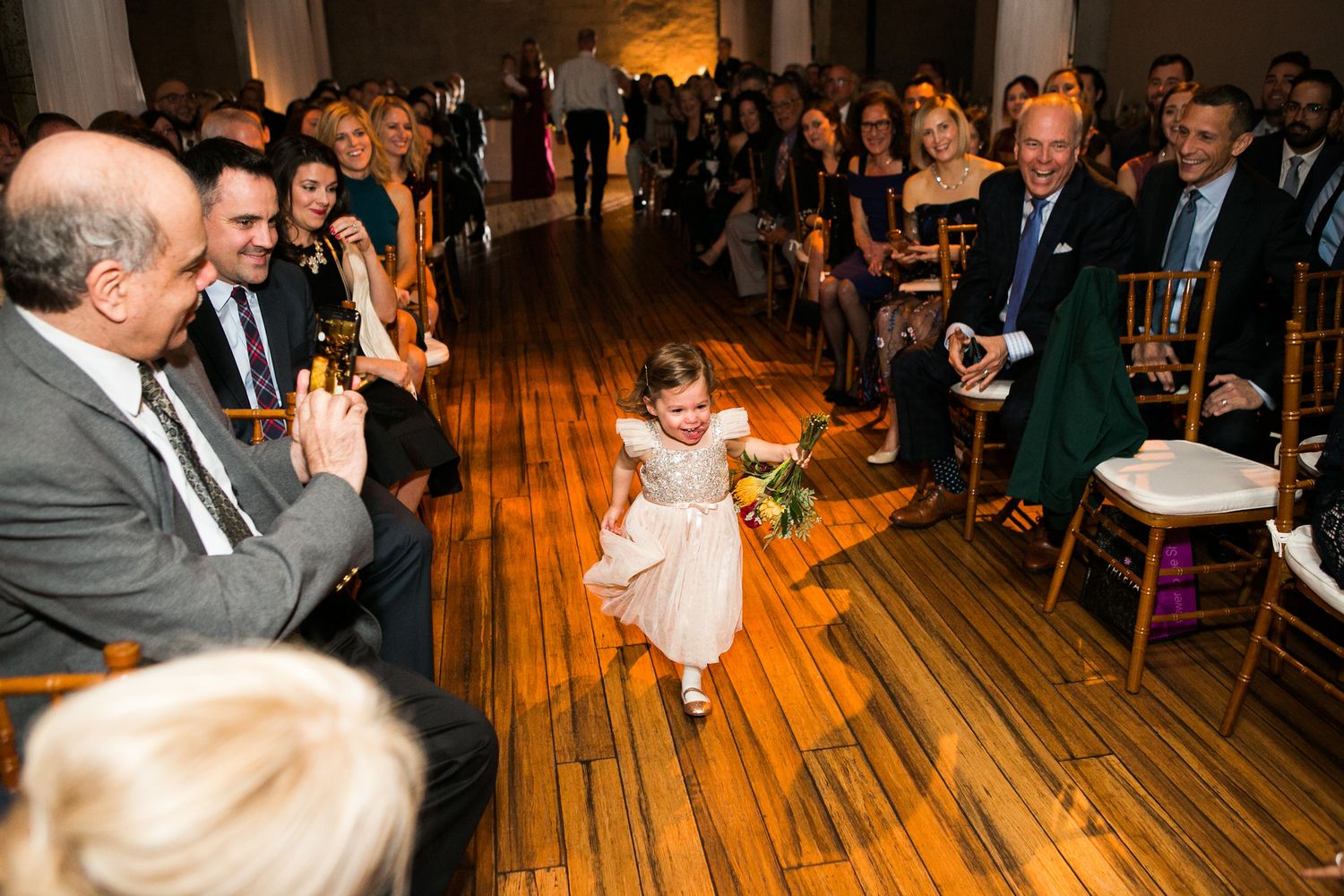 Adorable flower girl in white dress runs down wedding aisle as guests smile and applaud.