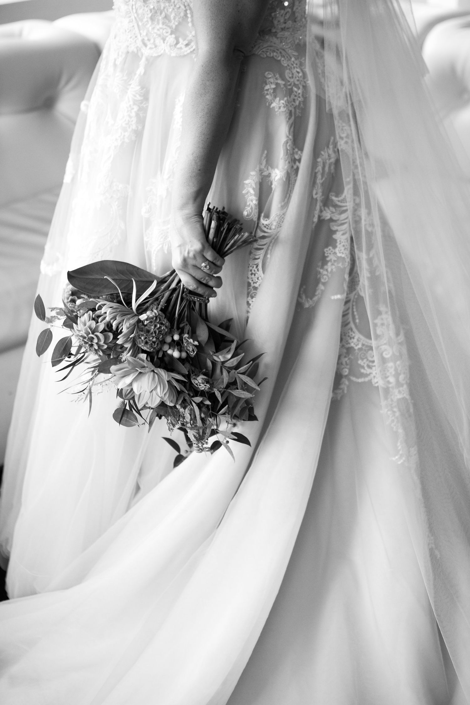 Black and white photo of bride in lace wedding gown holding floral bouquet with flowing veil.