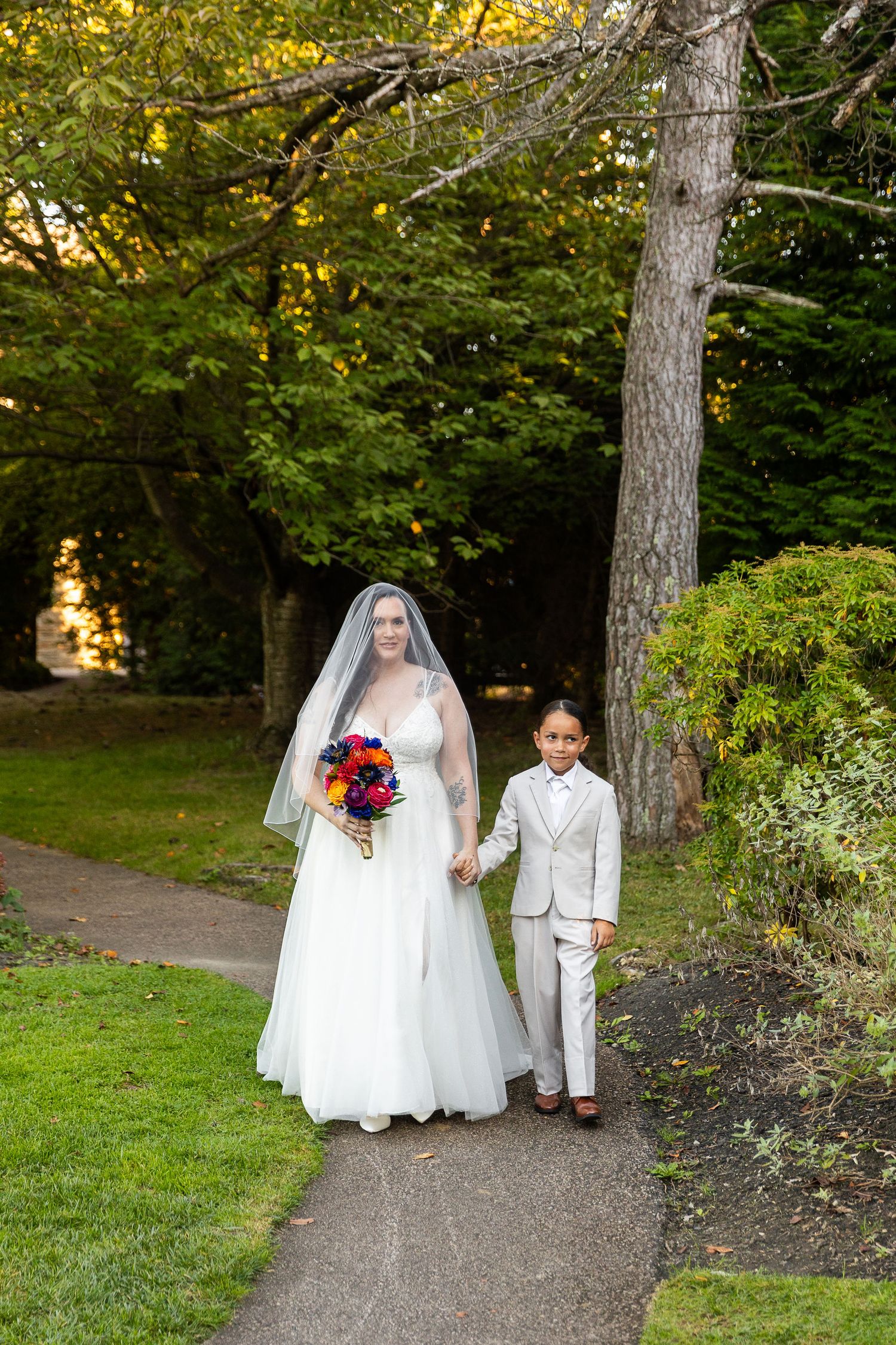 A bride in a white gown holding a colorful bouquet walks hand-in-hand with a young boy in a gray suit.