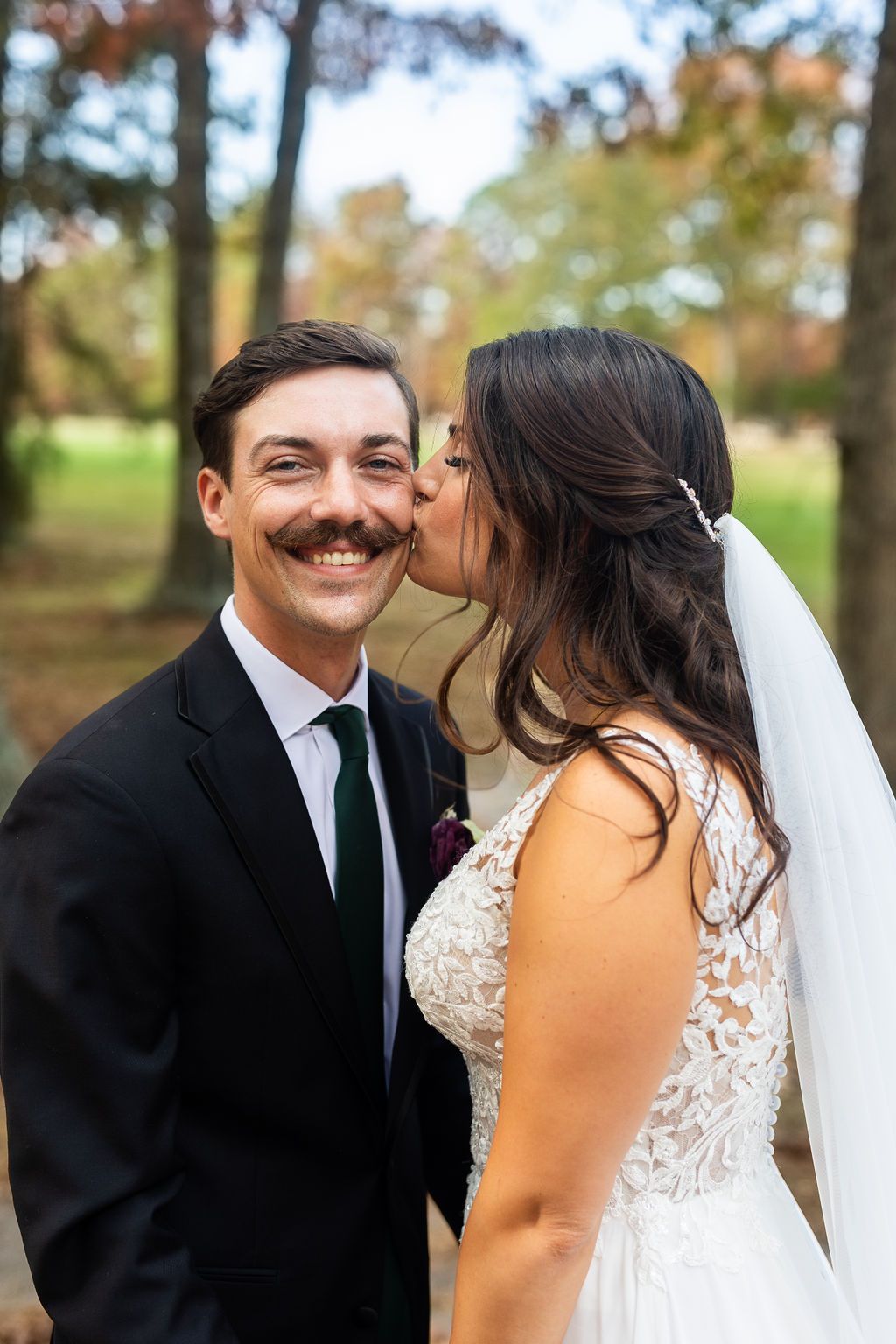 Bride in lace veil kisses smiling groom with mustache on cheek at fall outdoor wedding.