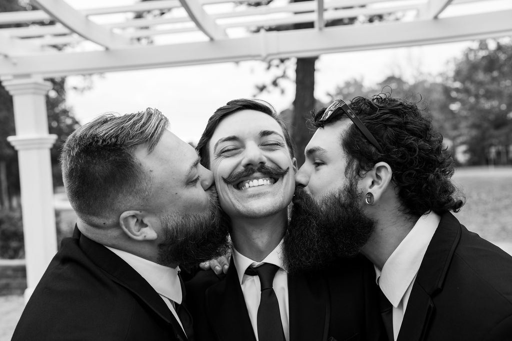 Black and white photo of two groomsmen kissing a laughing groom on both cheeks at wedding.