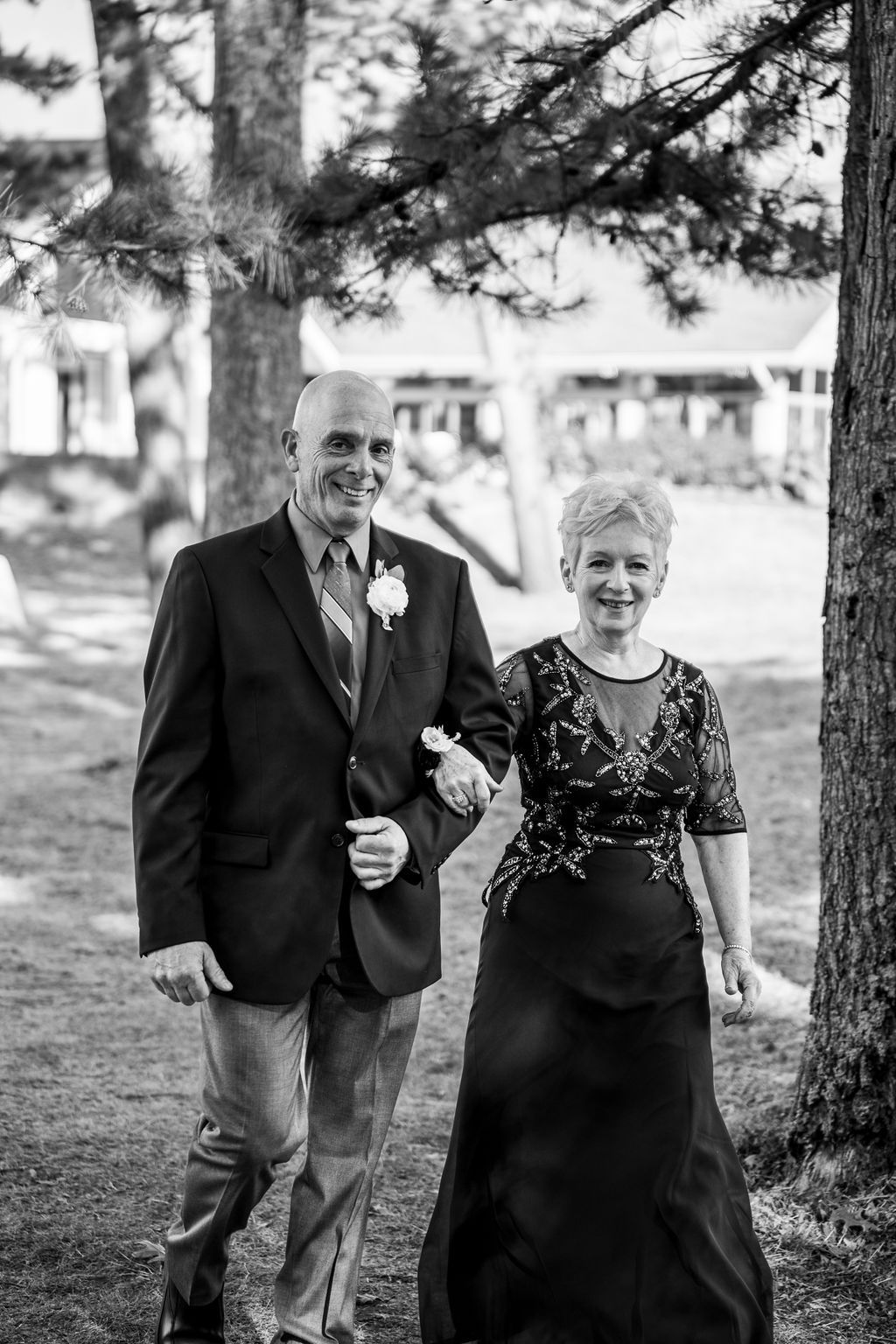 Black and white photo of smiling couple walking together at outdoor wedding, man wearing boutonniere, woman in elegant dress.