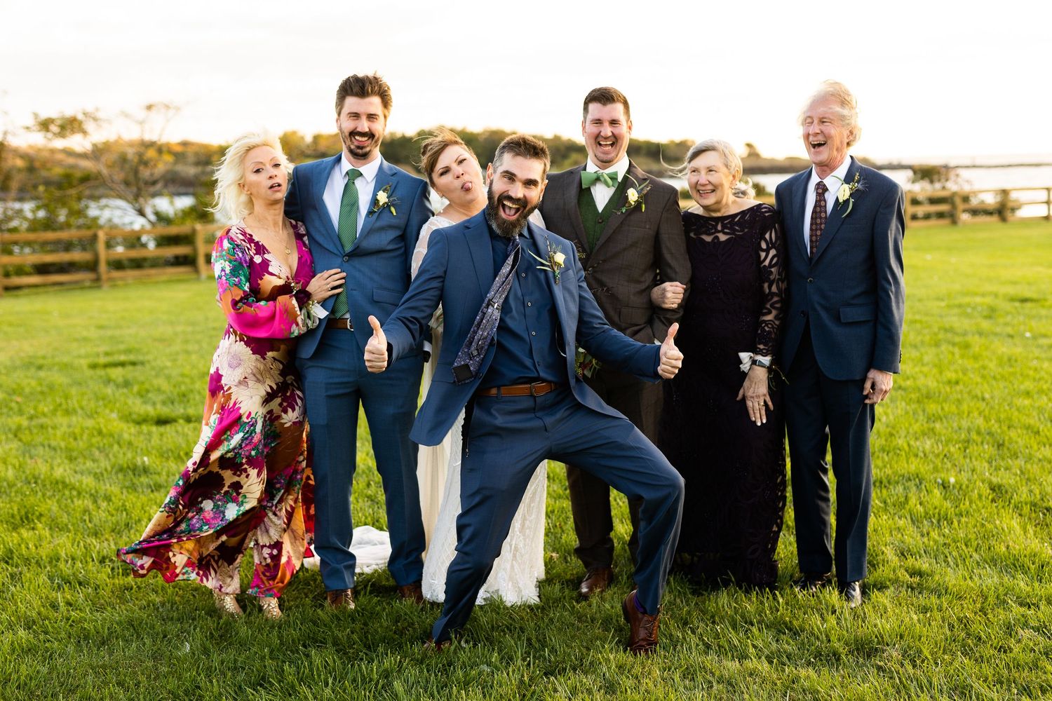 Joyful wedding party laughs and poses together on a green lawn at a waterfront venue.