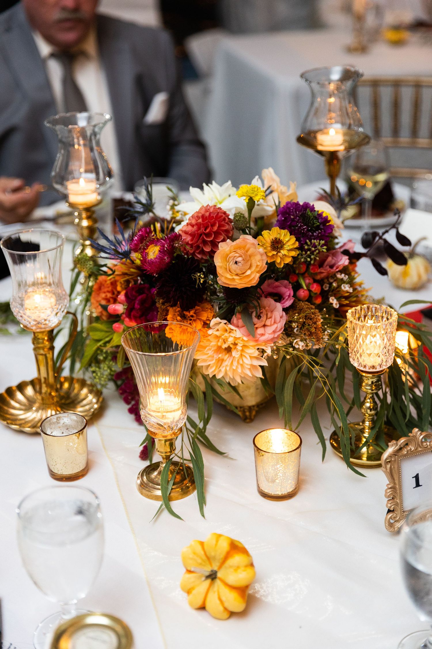 Fall wedding centerpiece with colorful flowers, gold candleholders, and a decorative mini pumpkin.