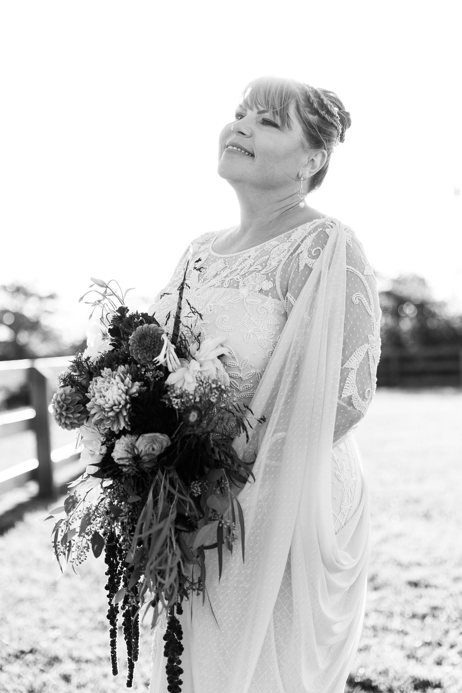 Bride in lace gown with draped veil smiling upward while holding cascading floral bouquet, black and white.