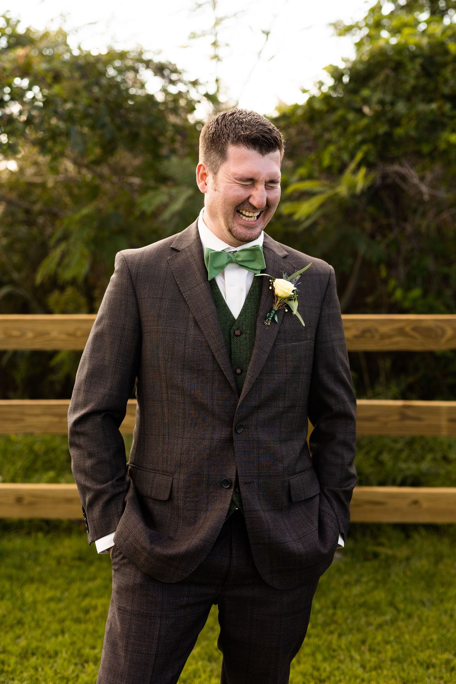 Joyful groom laughing in plaid brown suit with green bow tie and boutonniere at outdoor wedding.