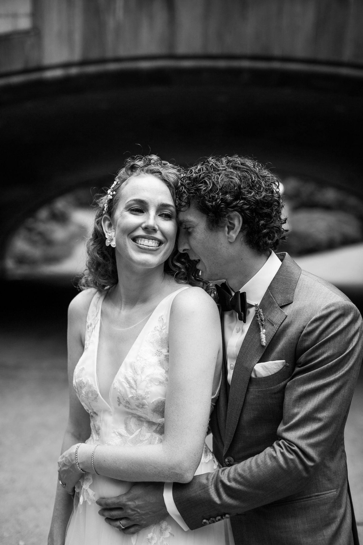 Black and white photo of groom whispering to laughing bride under a stone arch bridge on their wedding day.