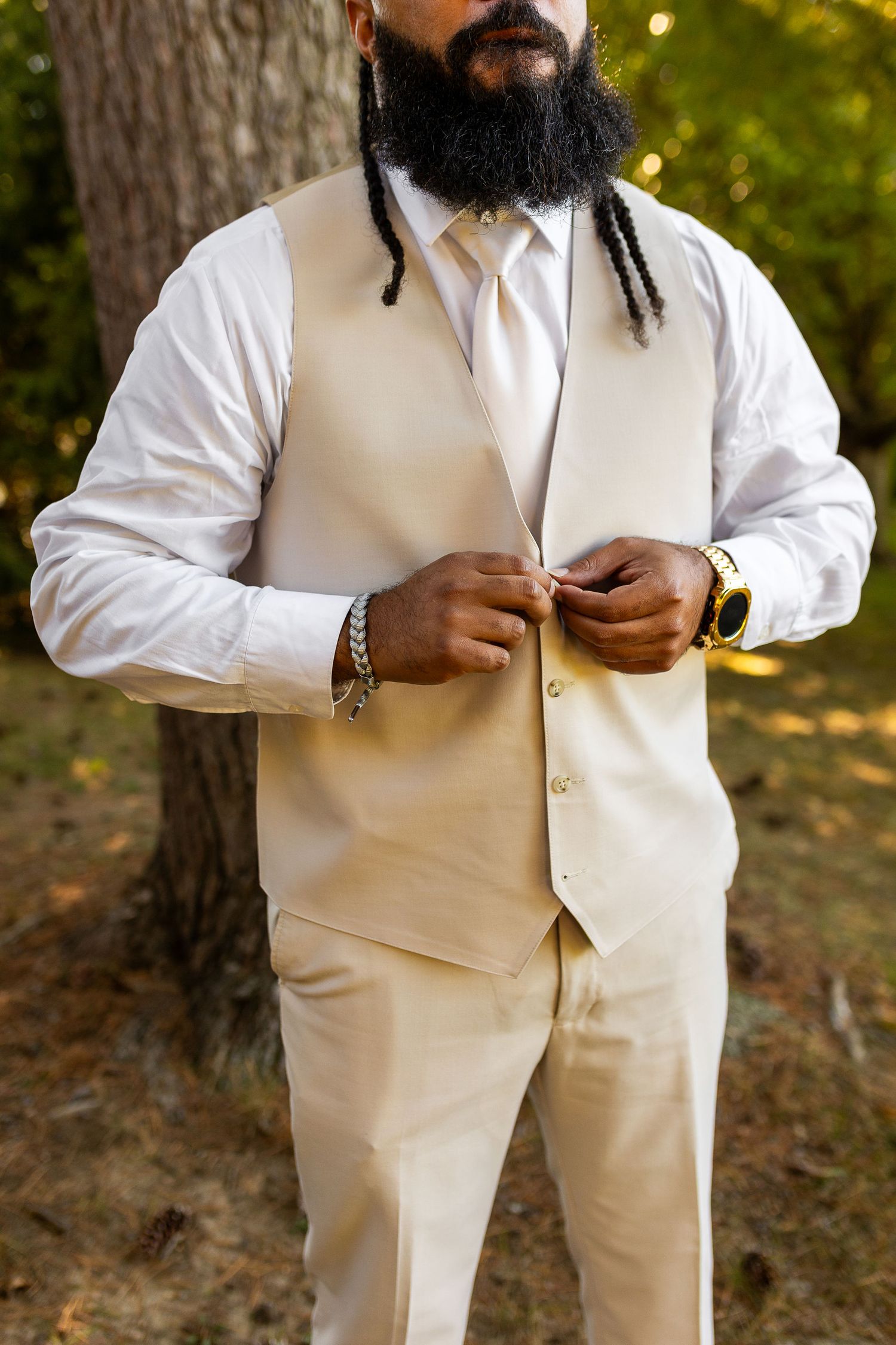 Groom with dreadlocks and beard in a cream wedding vest suit buttoning up near a tree outdoors.