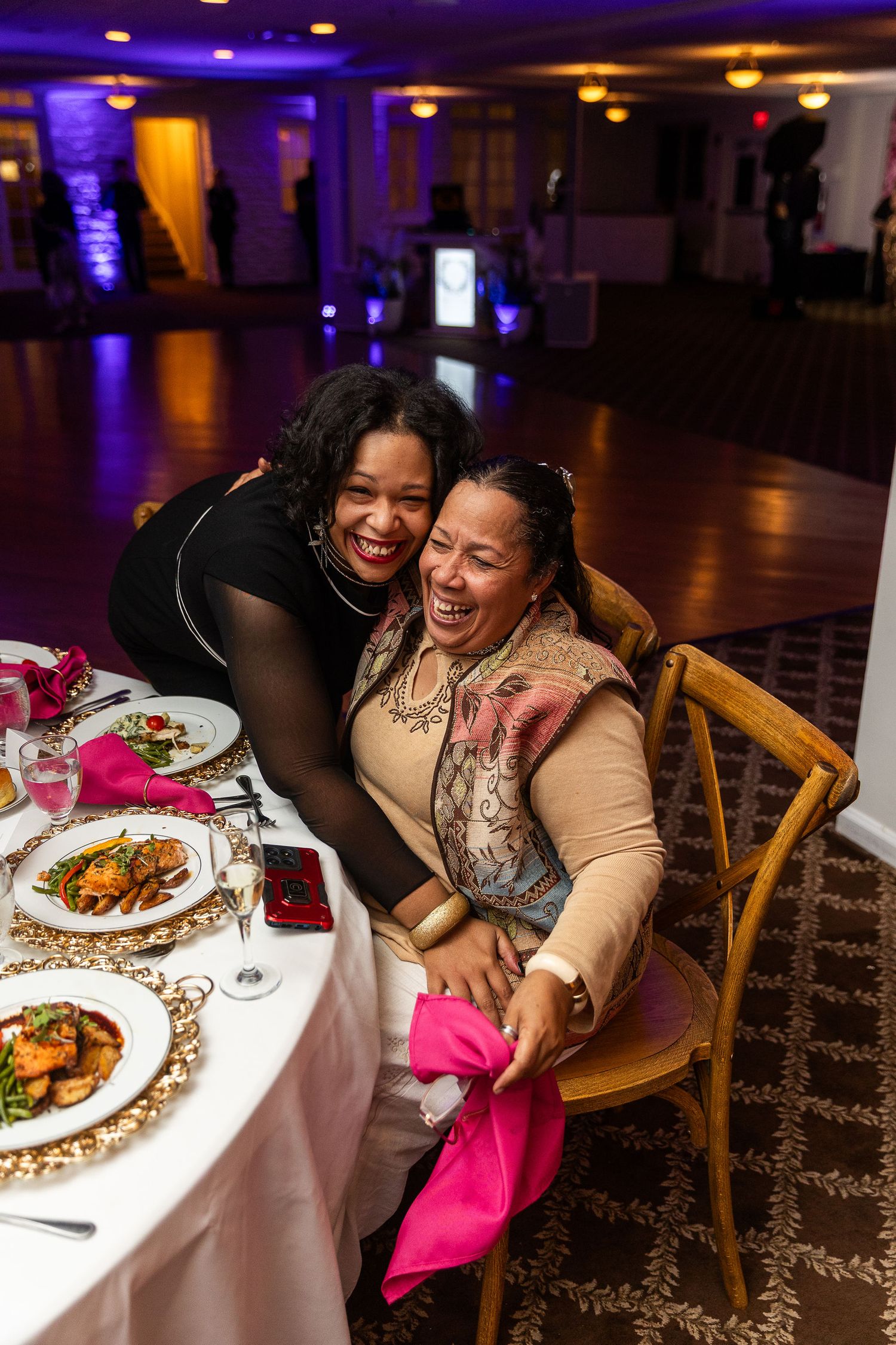 Two women laugh joyfully together at a wedding reception dinner table with pink napkins and elegant place settings.