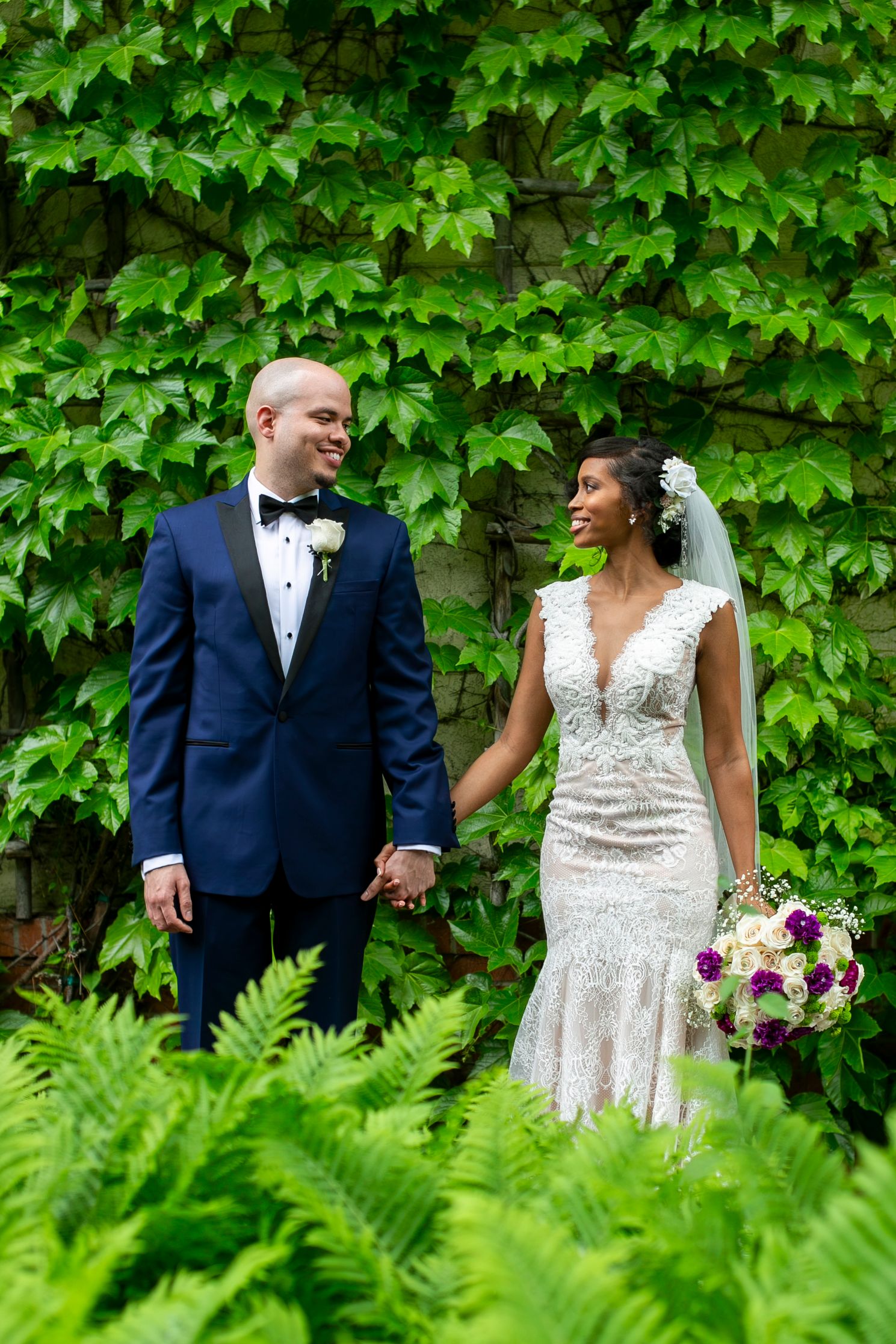 Interracial wedding couple holds hands smiling among green ferns and ivy-covered stone wall backdrop.