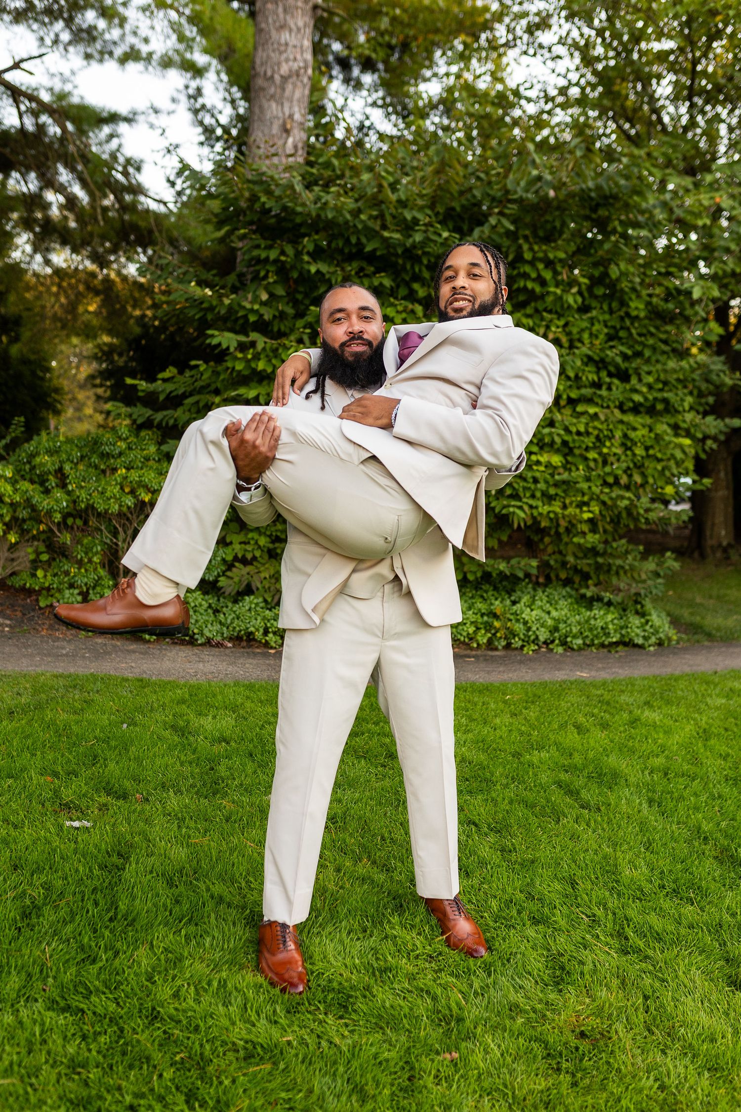 Two groomsmen in white suits laugh as one carries the other in a playful pose on a green lawn.