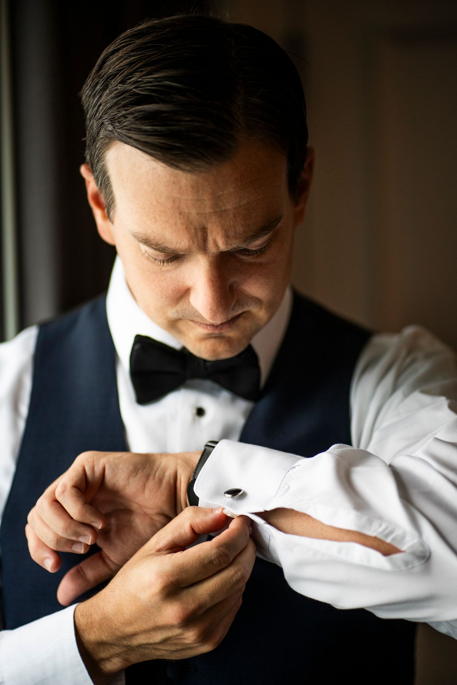 Dapper groom in navy vest and bow tie carefully fastening his cufflinks before the wedding ceremony.