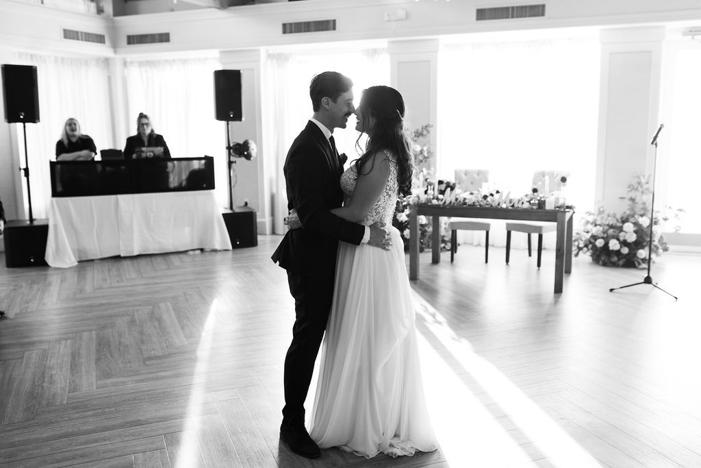 Bride and groom share intimate first dance in black and white photo with DJ setup visible in background.
