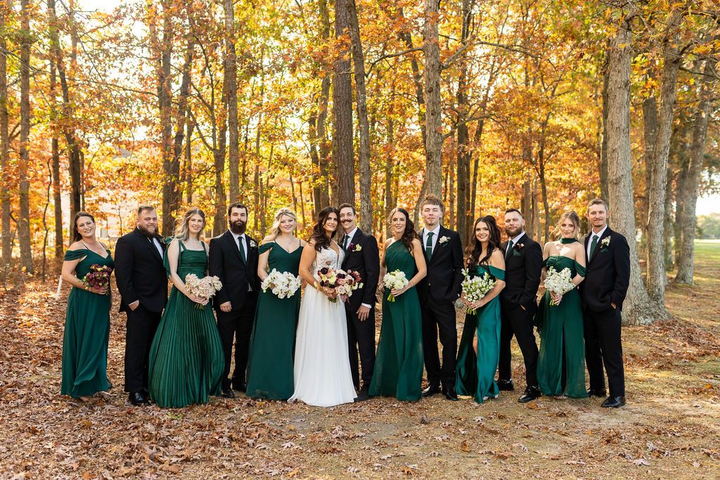 Full wedding party posing in emerald green and black attire among stunning autumn foliage in a forest.