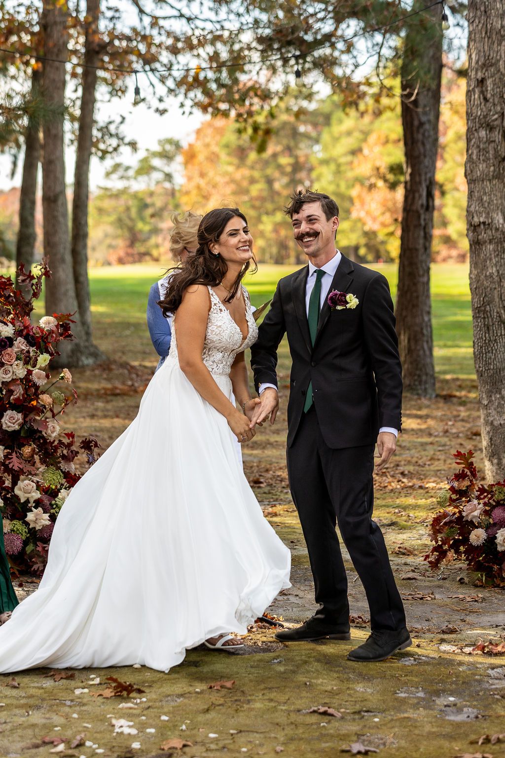 Joyful newlywed couple walks hand in hand after outdoor fall wedding ceremony surrounded by autumn foliage.